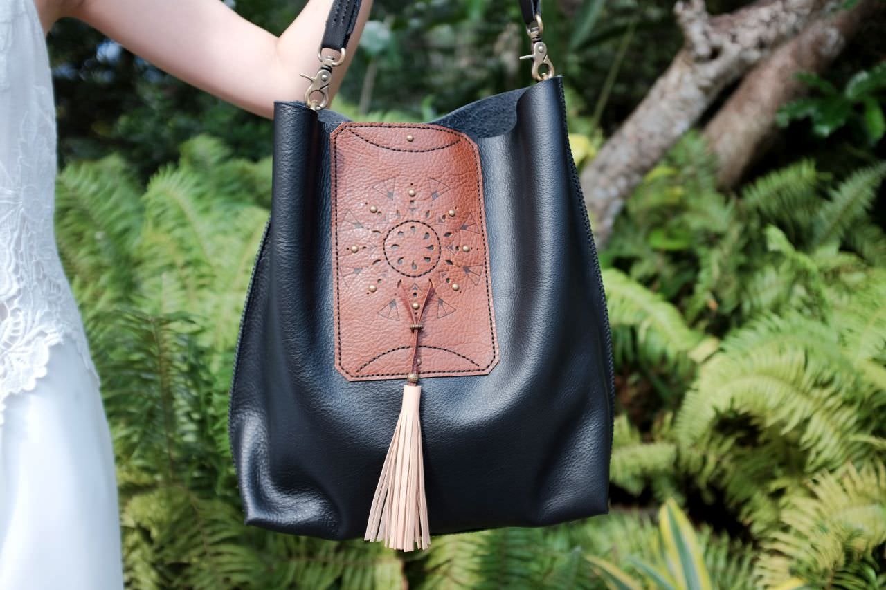 A black heather tote bag with a brown decorative panel and tassel, held by an anonymous hand in front of a green leafy backdrop, a product photo of a Vietnamese-made bag by leatherware producer Freewill Leather