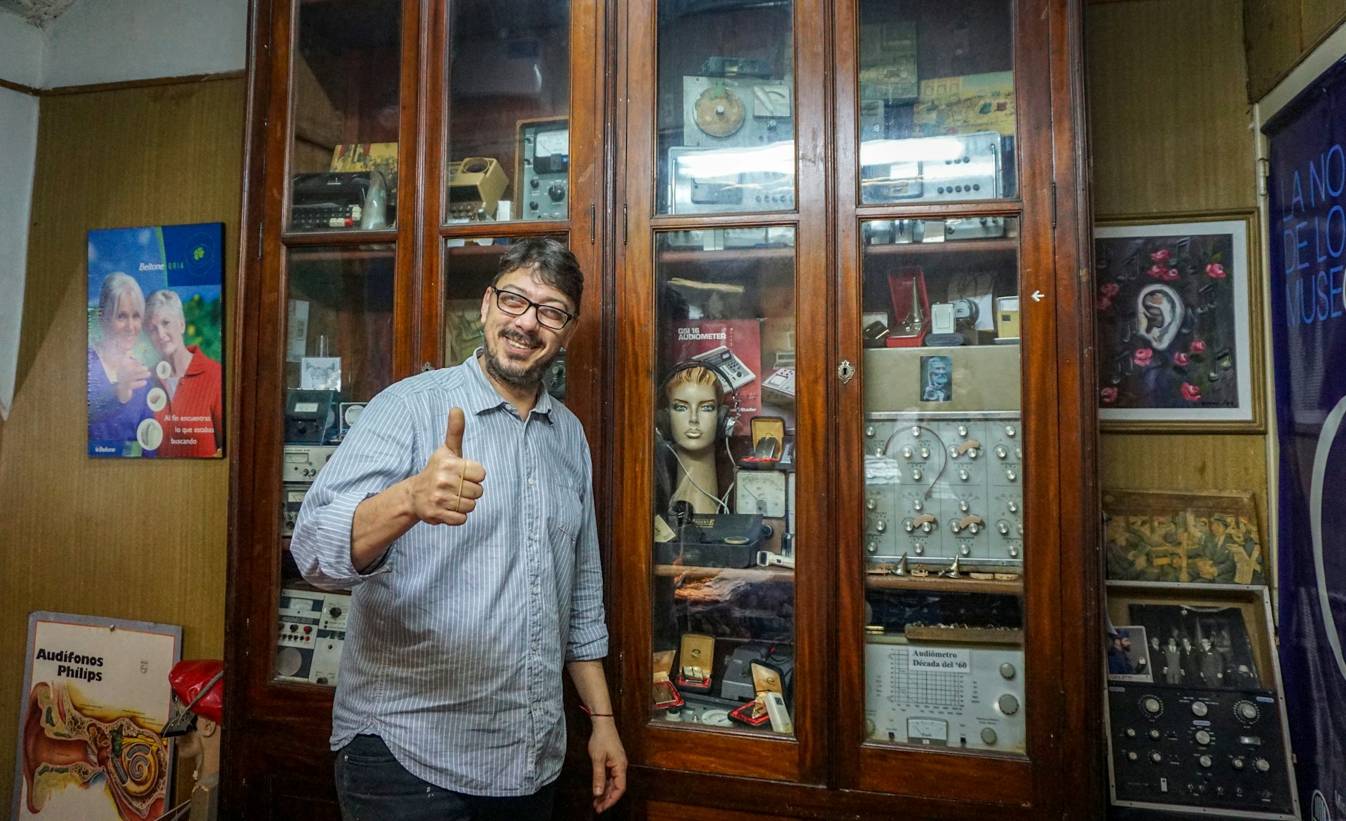 A man in a blue striped shirt and glasses giving the thumbs up stands in front of a cabinet full of hearing aid technology