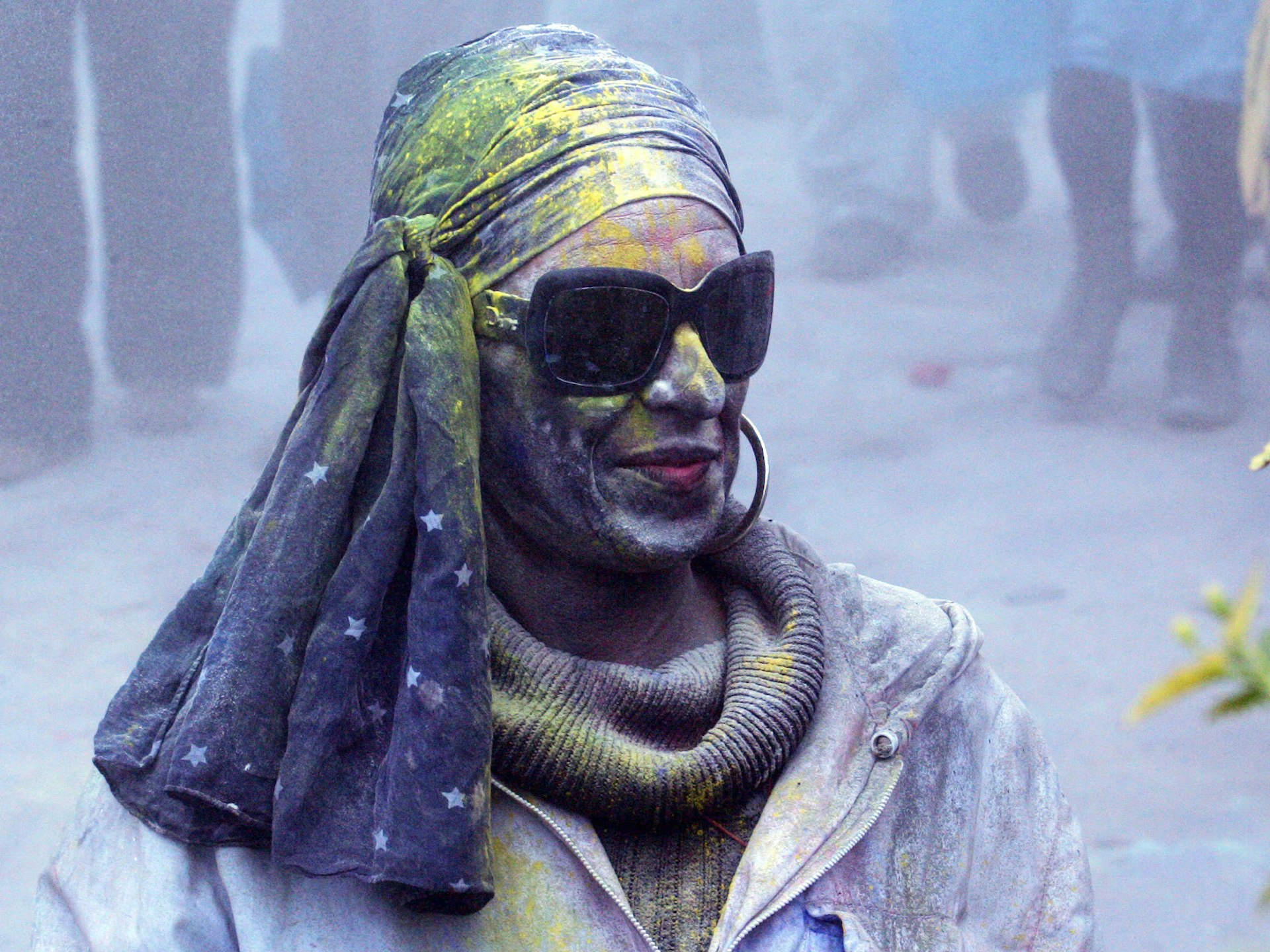 A person, wearing sunglasses and a cloth on their head, is covered in coloured pigment