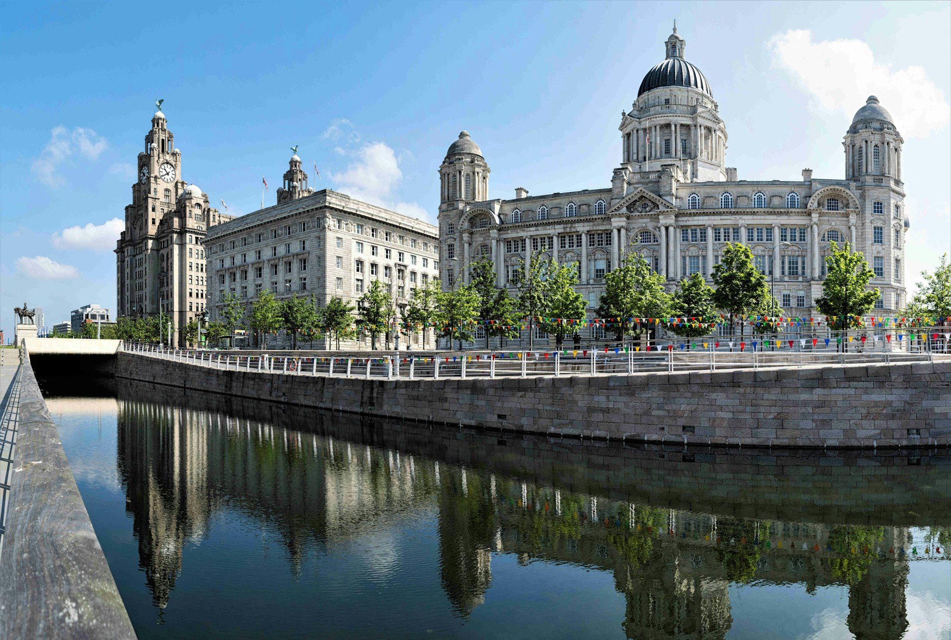 The Three Graces, Liverpool. The grey-stone buildings are reflected in the river water below and lined with luscious green trees as well as colourful bunting. 
