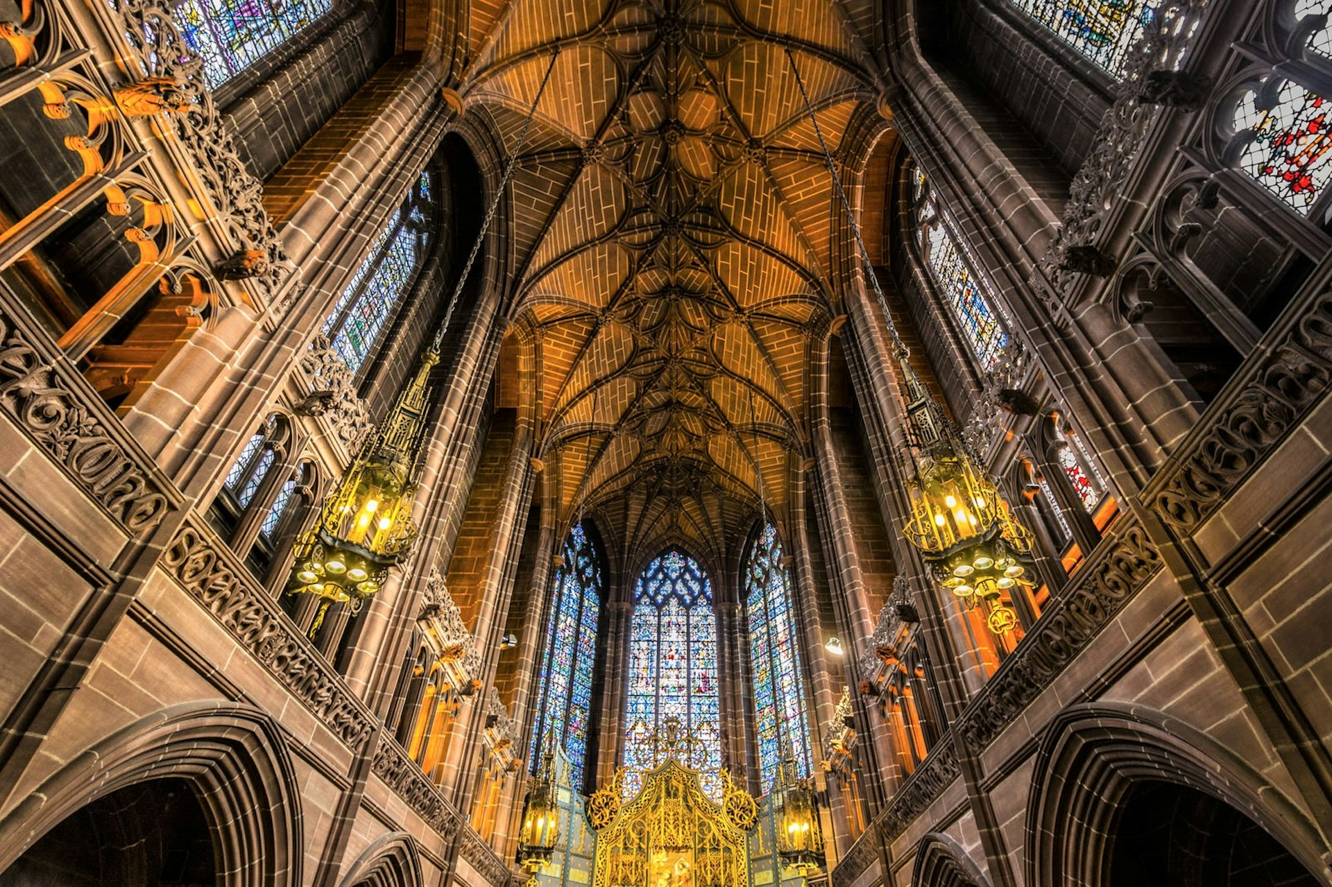 Liverpool's Anglican Cathedral's gothic roof is lit by yellow-hued lamps. The colourful stained-glass windows are a dramatic feature. 