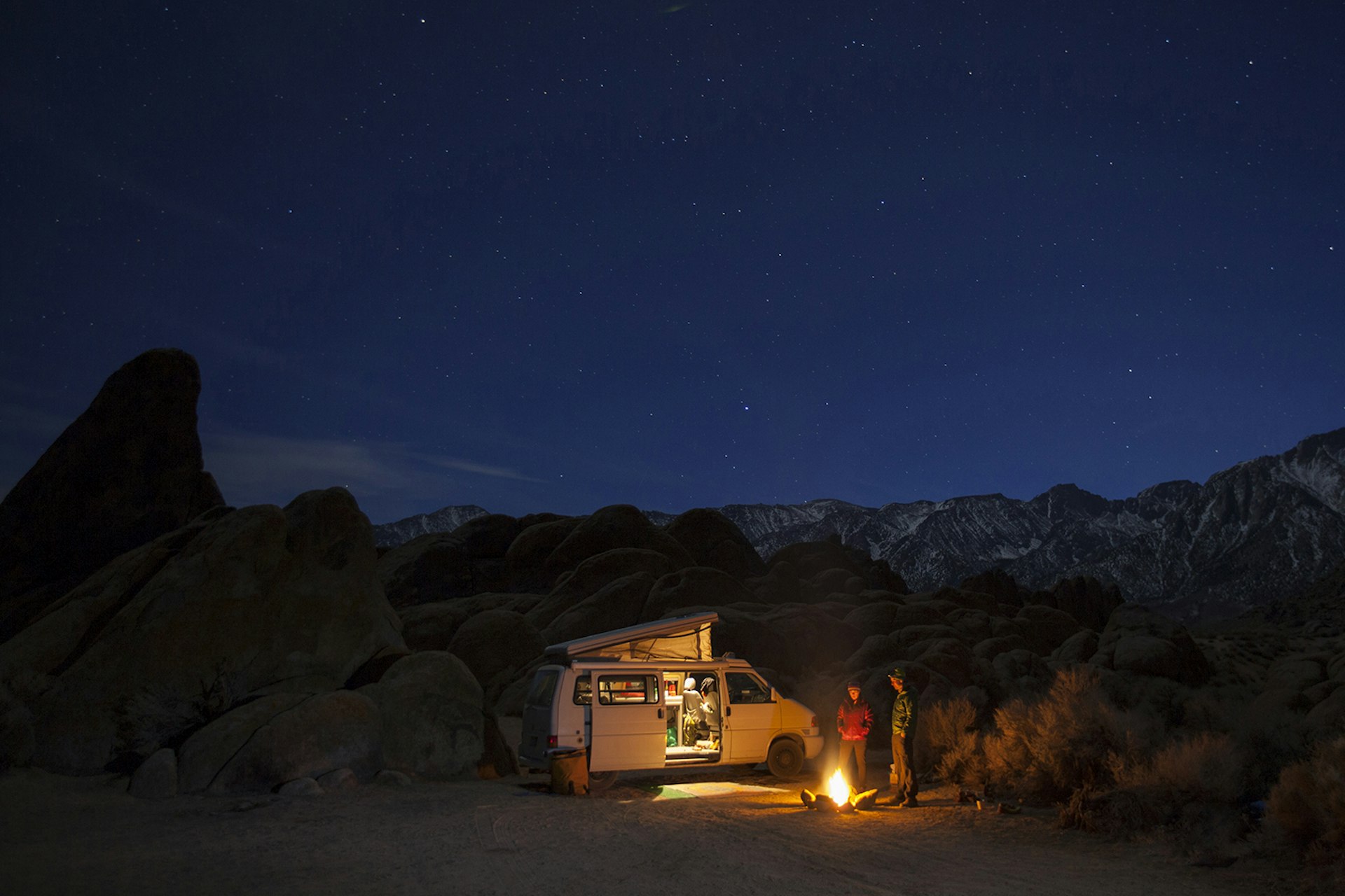 Two people standing by a campfire outside their campervan