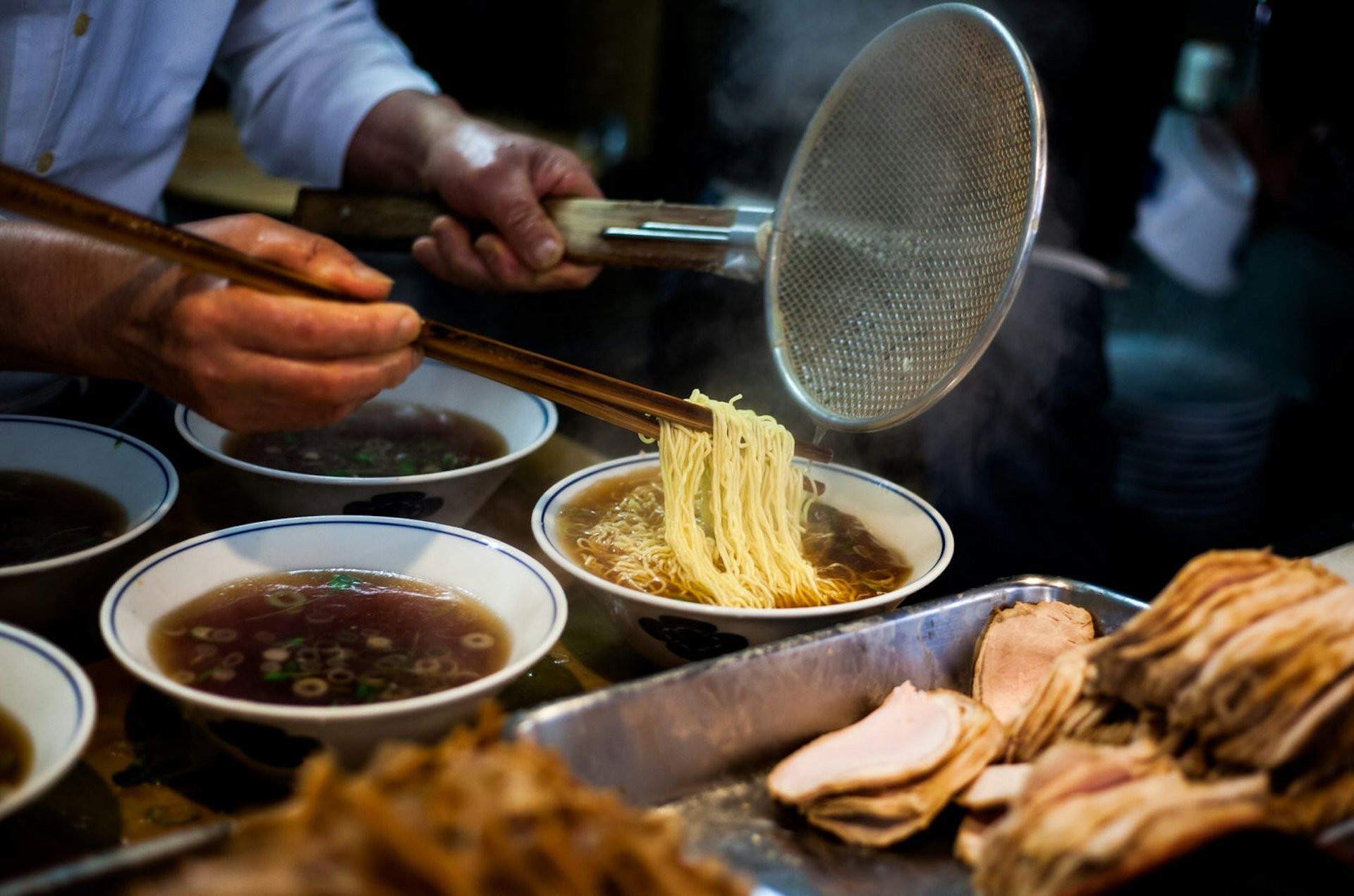 Hands preparing steaming ramen (soba) while holding long chopsticks and metal strainer. Several bowls of broth, and a metal tray of sliced pork, are in the foreground.