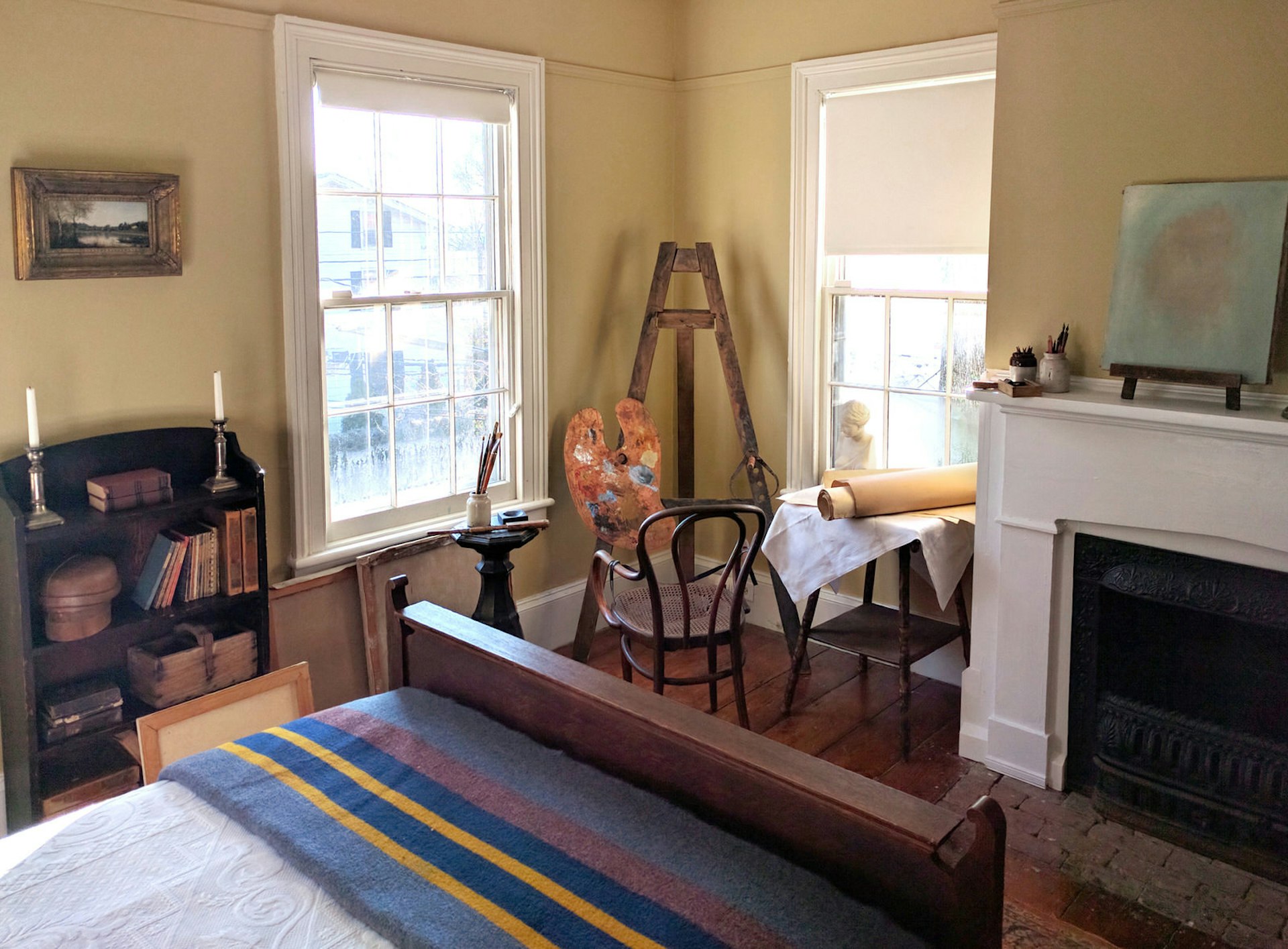 A sunny, yellow-walled room with a trim, well-made wooden bed and an unlit fireplace. A painter's easel is in the corner of the room. The room is brightly lit through two large windows. It's the bedroom at Edward Hopper House Museum in Nyack, New York