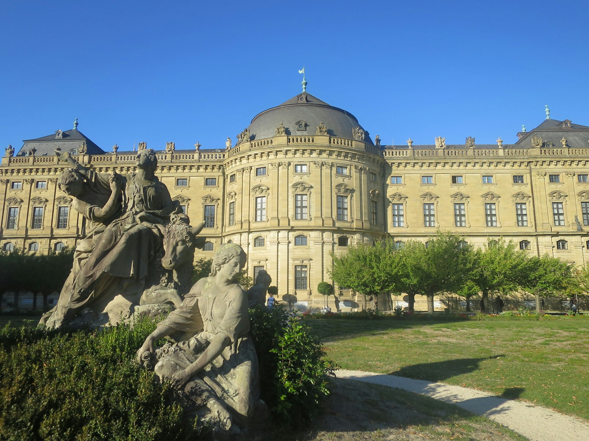 Würzburg Residenz, an impressive palace in Austrian/German baroque style, also inspired by Versailles. The palace lawn is adorned with trees and sculptures. its yellow facade contrasts with the bright blue sky