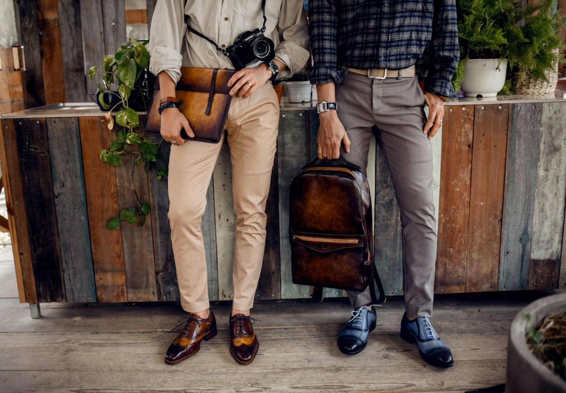 The legs and mid-sections of two men, each wearing neutral coloured slacks and white/checked shirts, and fine leather shoes. Both men carry polished leather bags in this product shot from leather purveyor Dominique Saint Paul, a Vietnam-based designer.