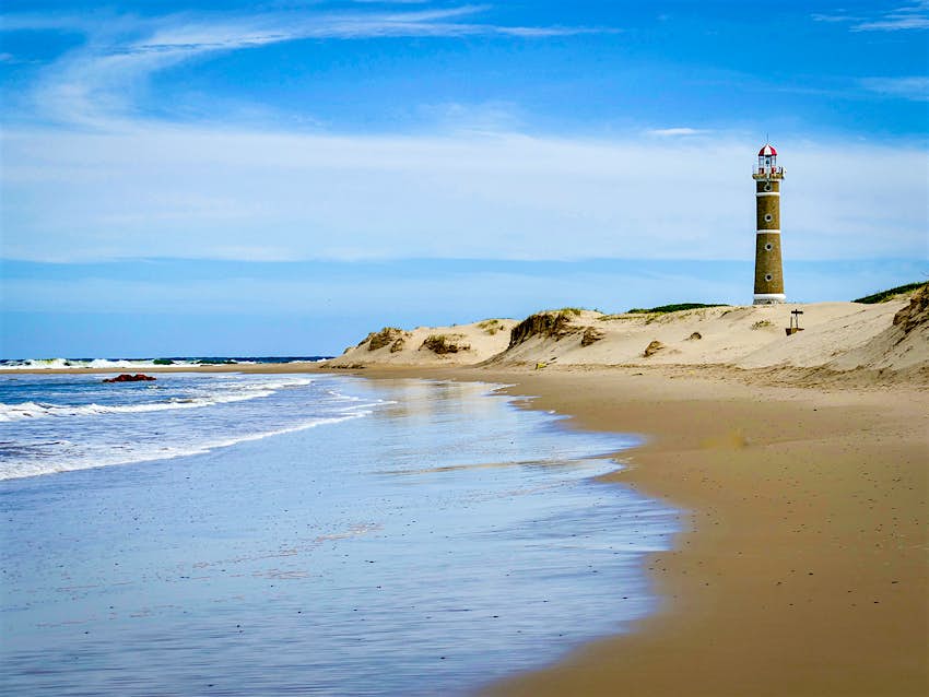 An empty beach with gold sand on a sunny day, with a lighthouse in the background on the right