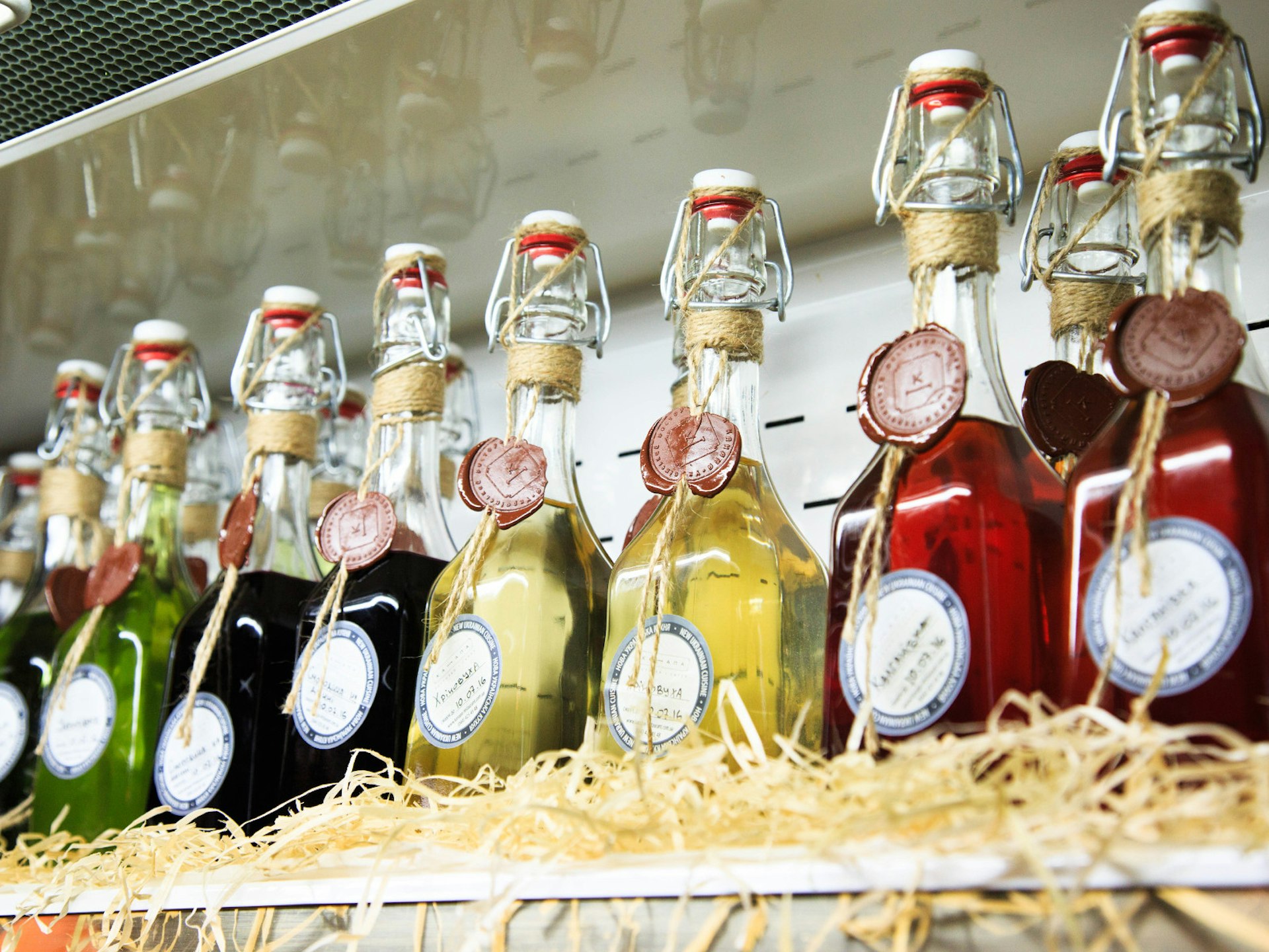 Close up of a row of colorful bottles on a straw-lined shelf, each full of a homemade ratafia or balm.