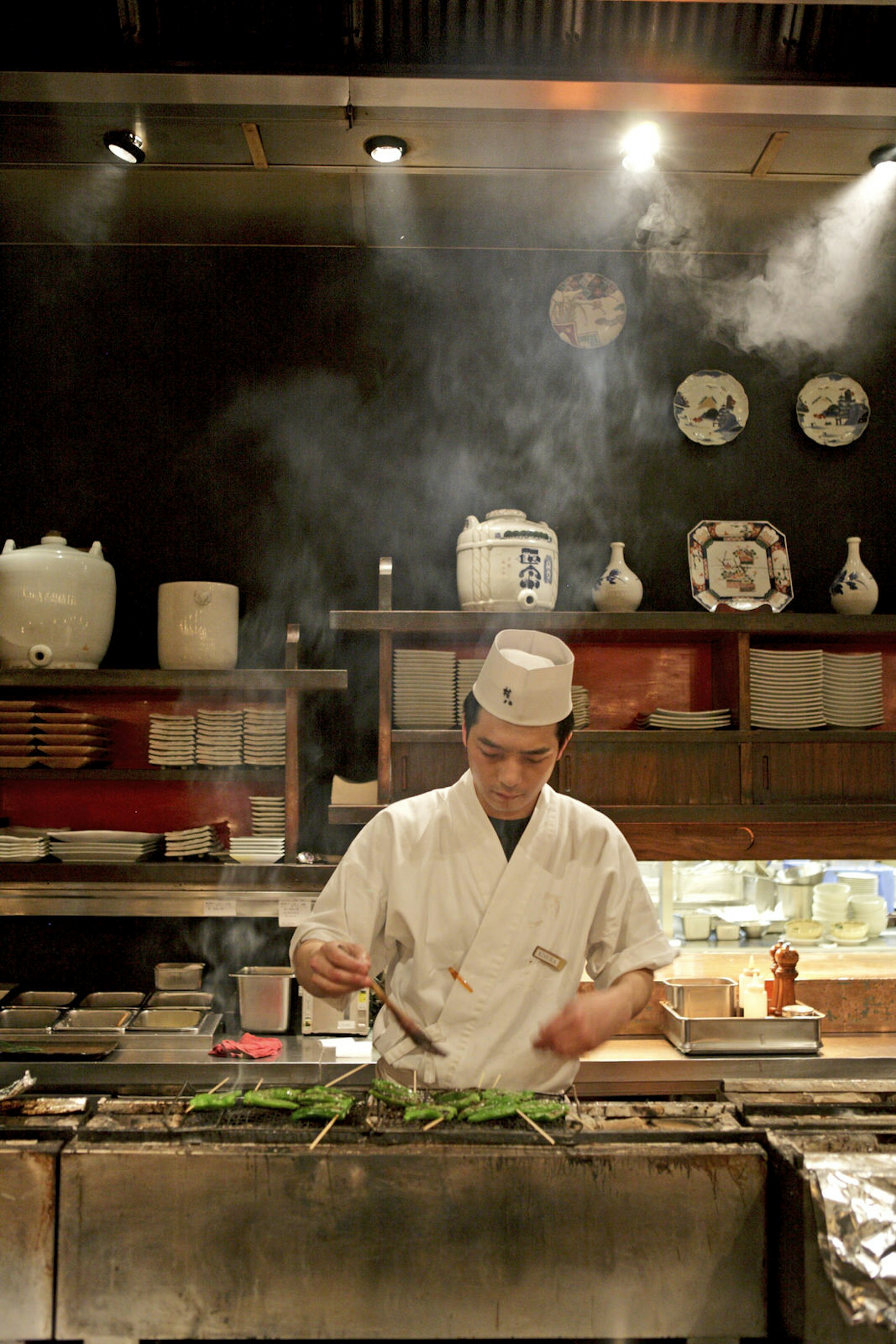 A white-clad chef wearing a hat delicately paints a dressing on some greens that are atop a smoking grills. At the back of this open kitchen, at a restaurant in Tokyo's Roppongi district, are shelved with white ceramic bowls and kitchenware.