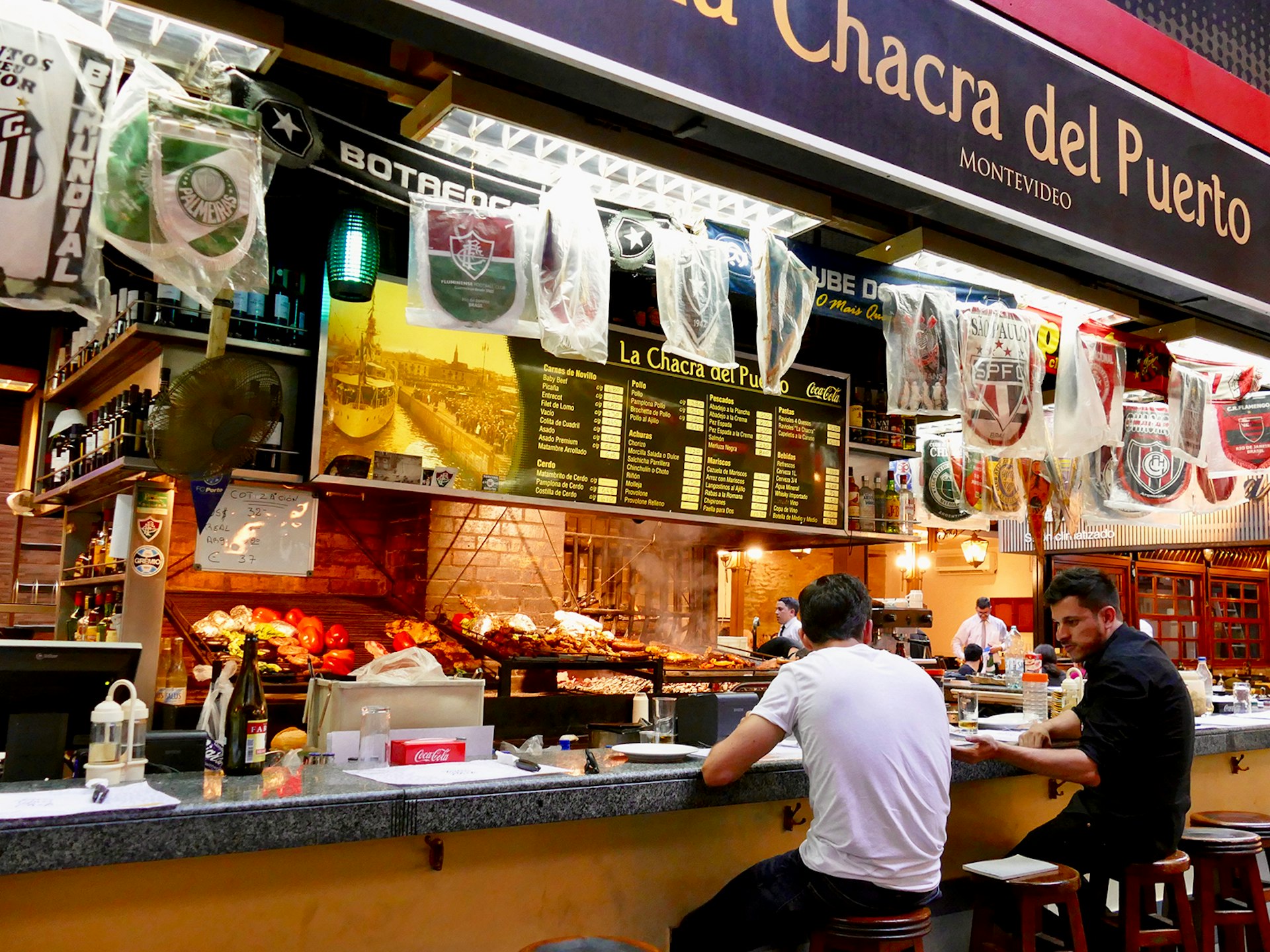 Two men sit at a colorful lunch counter in a market, with a menu on the wall in front of them above several grills