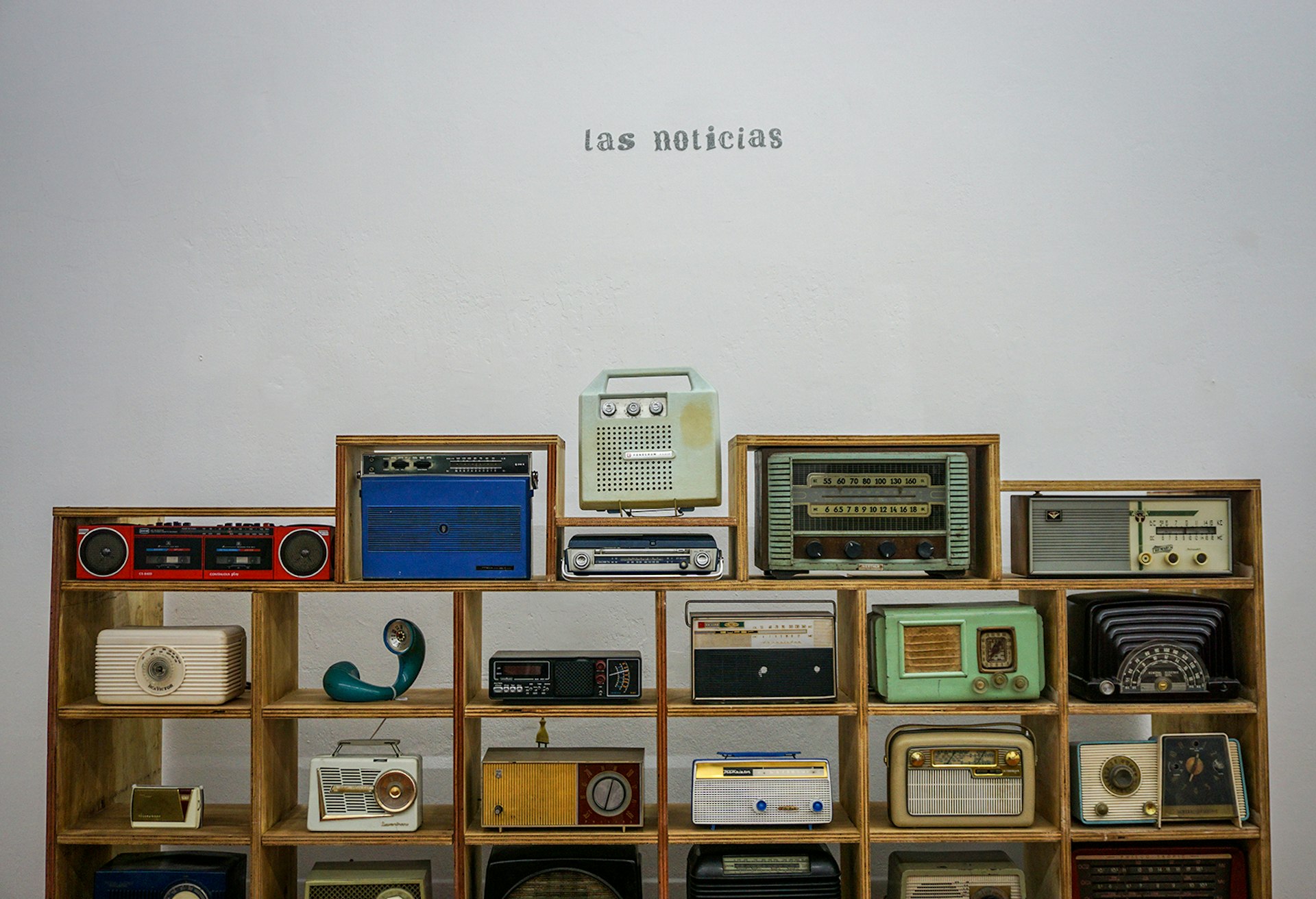 Old radios stacked in a mid-century style shelf against a blank white wall, with "las noticias" printed on the wall above them