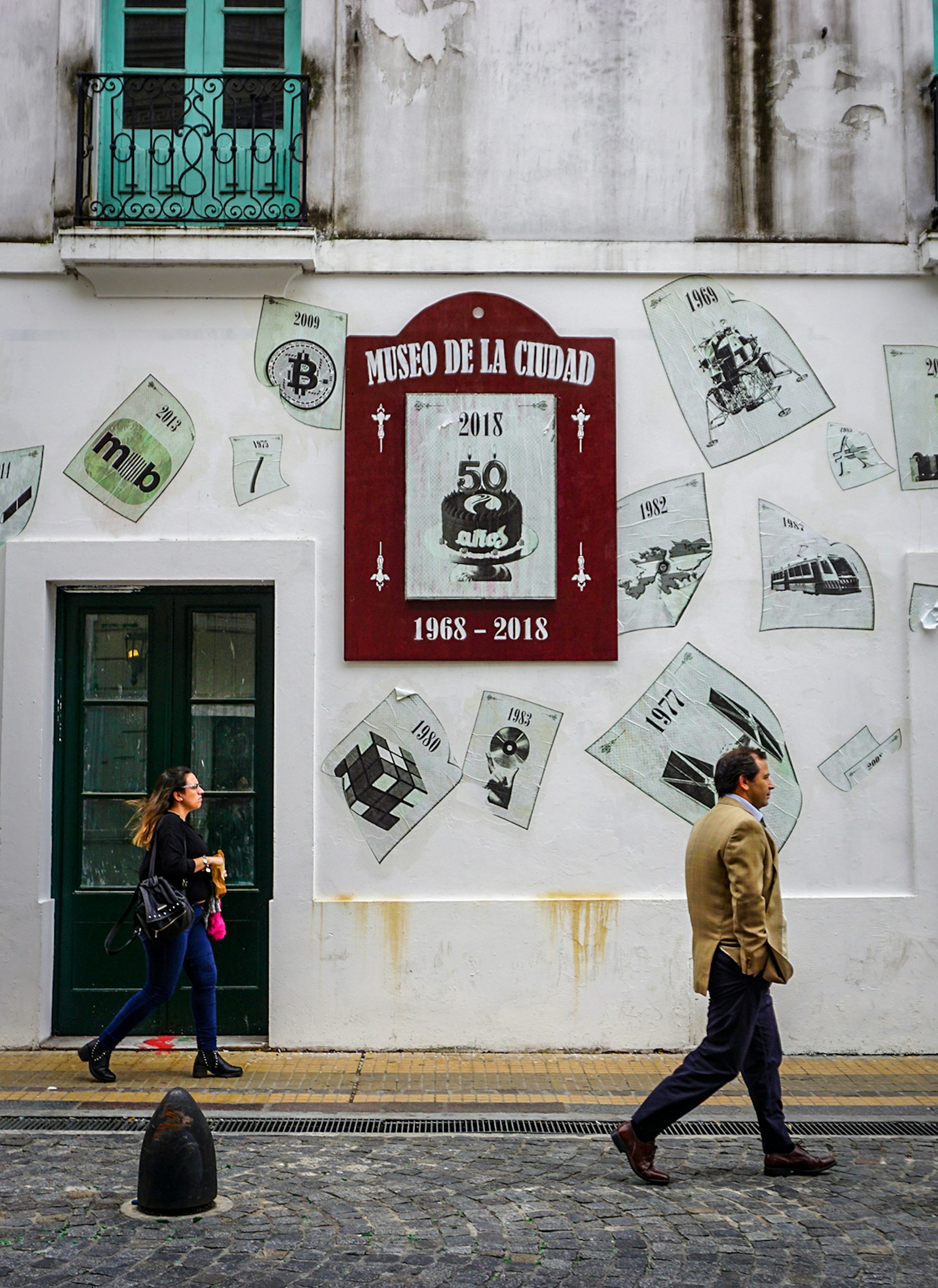 An exterior shot of two people walking past a red sign reading Museo de la Ciudad, which is pasted to a white, colonial building and surrounded by flyers depicting items you might see inside