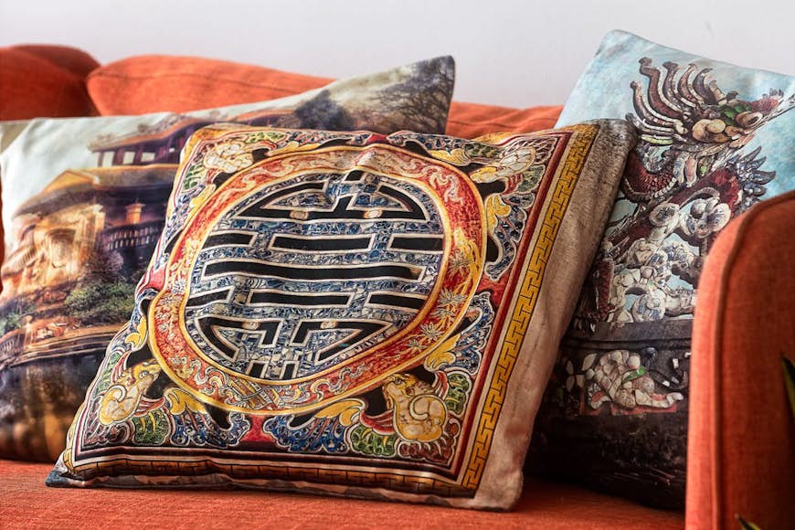 Två dekorativa kuddar, med slående geometriska mönster i vietnamesisk stil, vilar på en ockrafärgad soffa på denna produktbild, och illustrerar varorna som erbjuds av den vietnamesiska designern O&M