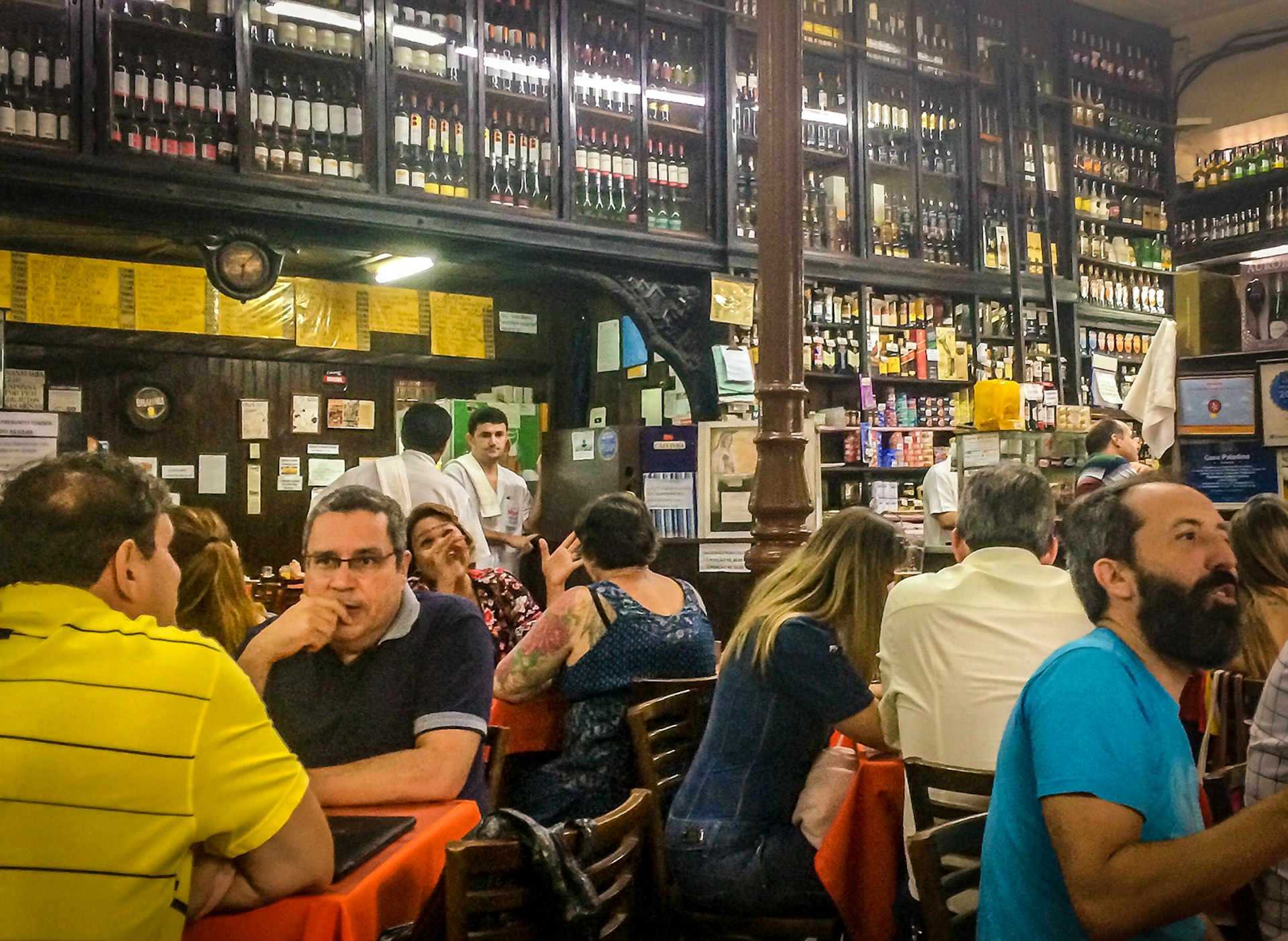 A crowded bar with a wall of dark wooden shelves lined with cachaca bottles