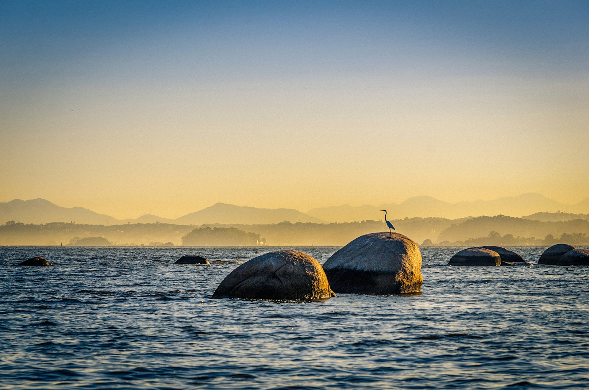 A view of the Guanabara Bay with round rocks poking up through the water's surface. A heron stands on the biggest rock.