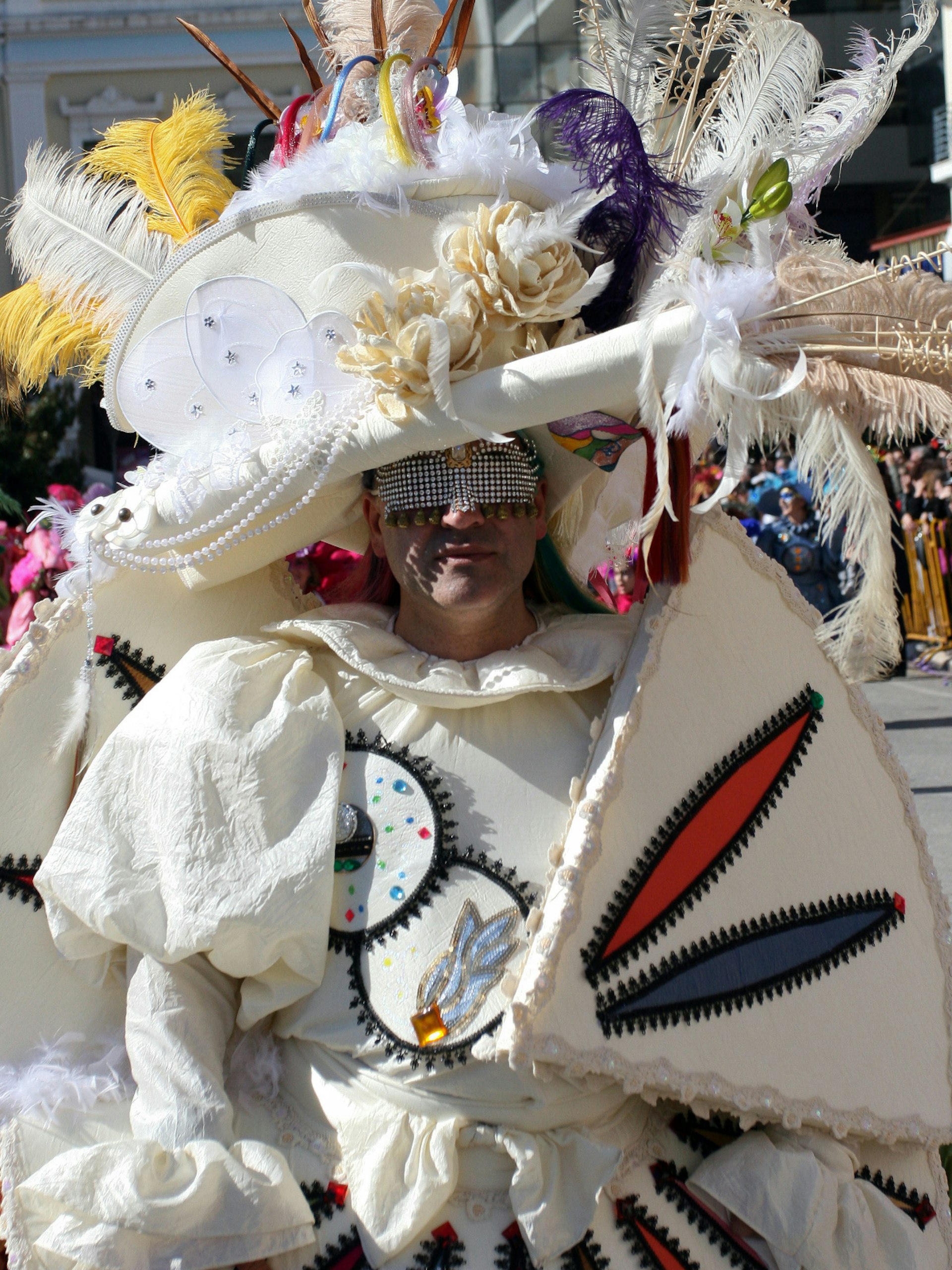 A person wearing a beaded mask is dressed in white with colourful patches, and elaborate head wear