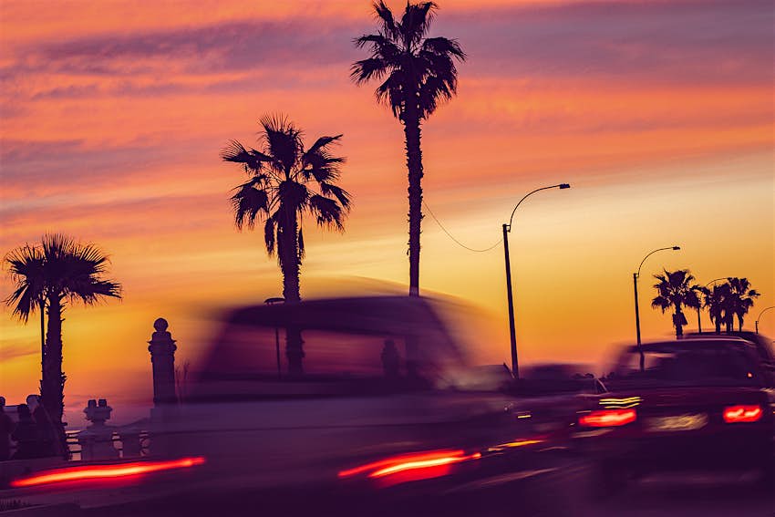 A long exposure of cars driving past palm trees, all silhouetted against a pink sunset sky