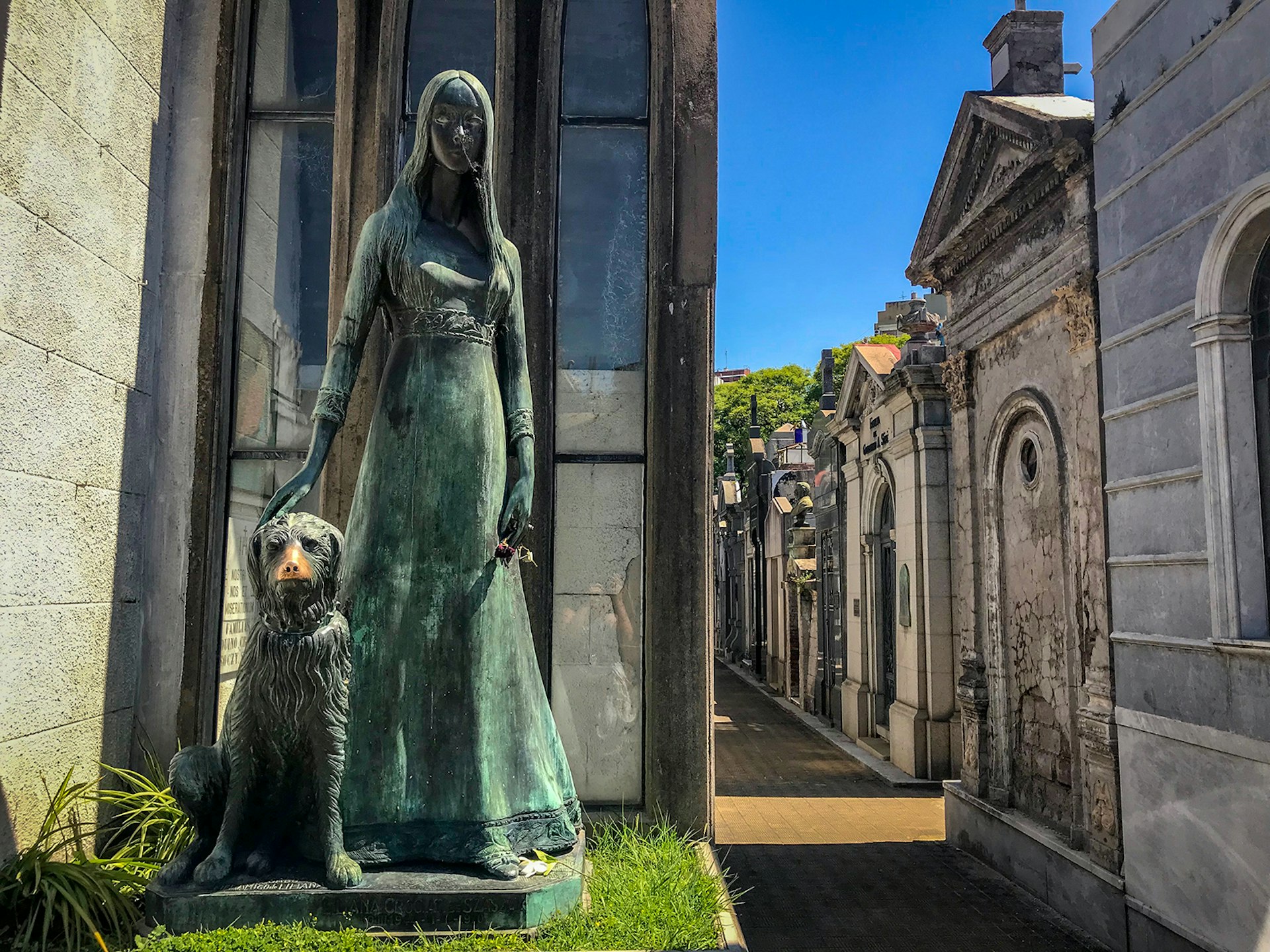 A statue of a woman with long hair petting a dog sitting at her side, with a row of mausoleums in the background