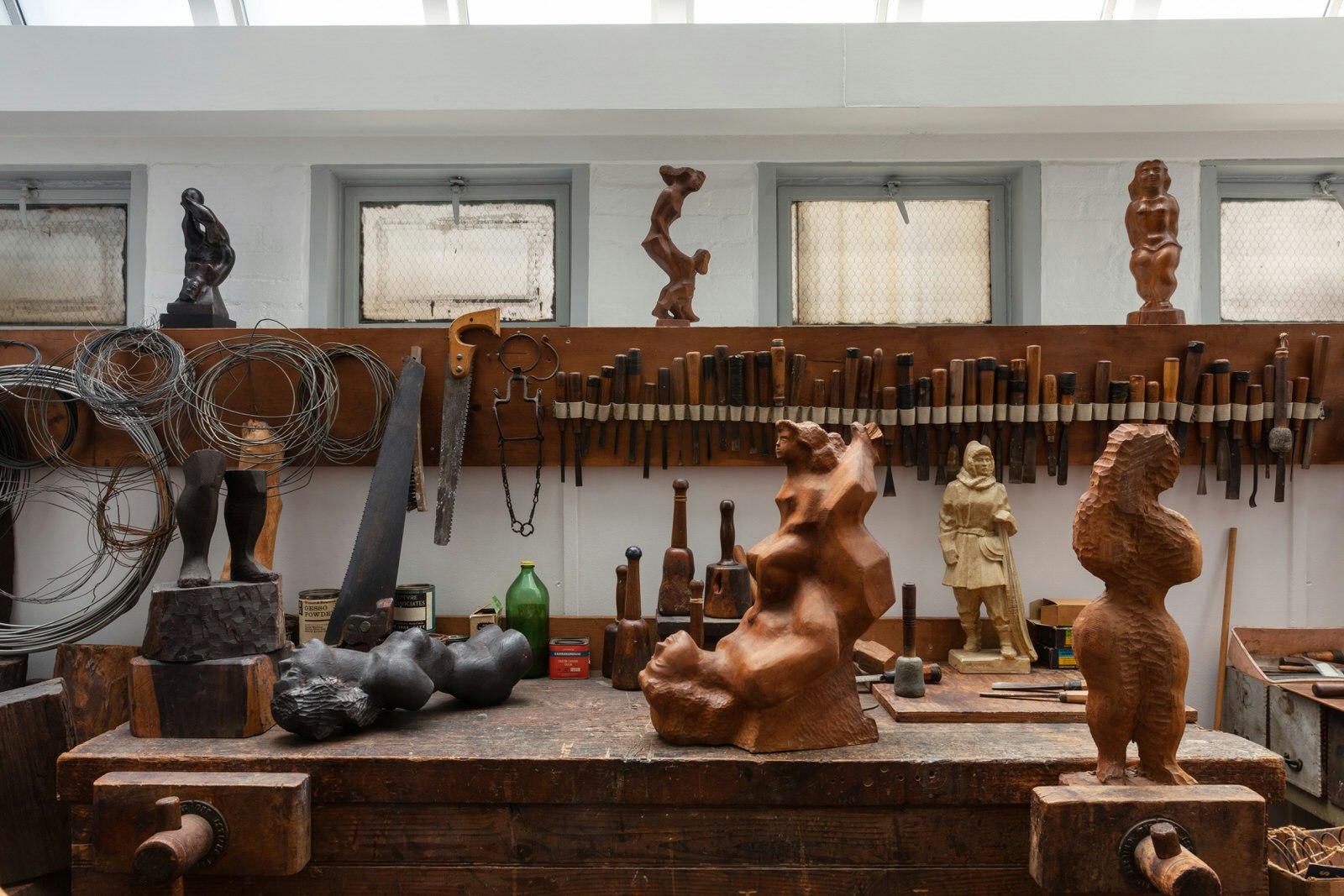 A wooden workbench covered in sculptures, with an array of different hand saws hooked onto the wall behind, a scene at the studio in Renee & Chaim Gross Foundation Museum