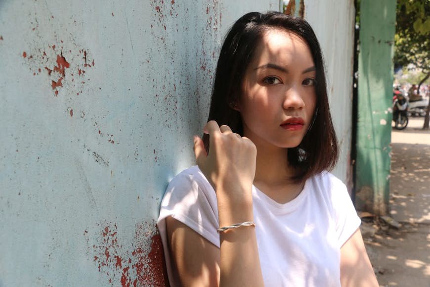 En ung mörkhårig kvinna med rött läppstift, med ett seriöst uttryck och klädd i en lös vit T-shirt, håller handleden mot kameran och modellerar ett enkelt men snyggt metallarmband, en av produkterna som säljs av Saigon Armory i Vietnam