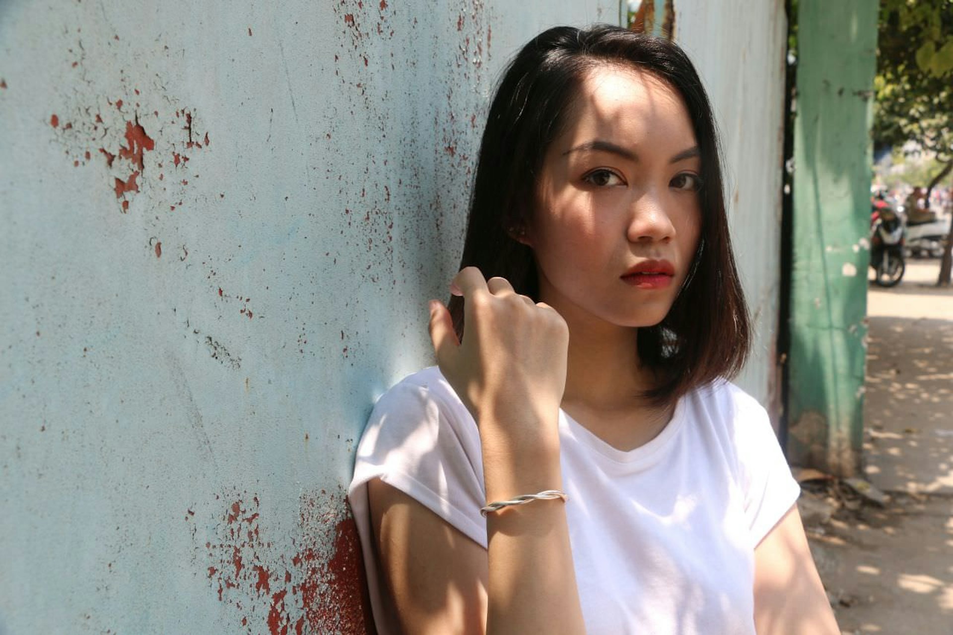 A young dark-haired woman with red lipstick, sporting a serious expression and wearing a loose white T-shirt, holds her wrist towards the camera, modelling a simple but stylish metal bracelet, one of the products sold by Saigon Armory in Vietnam