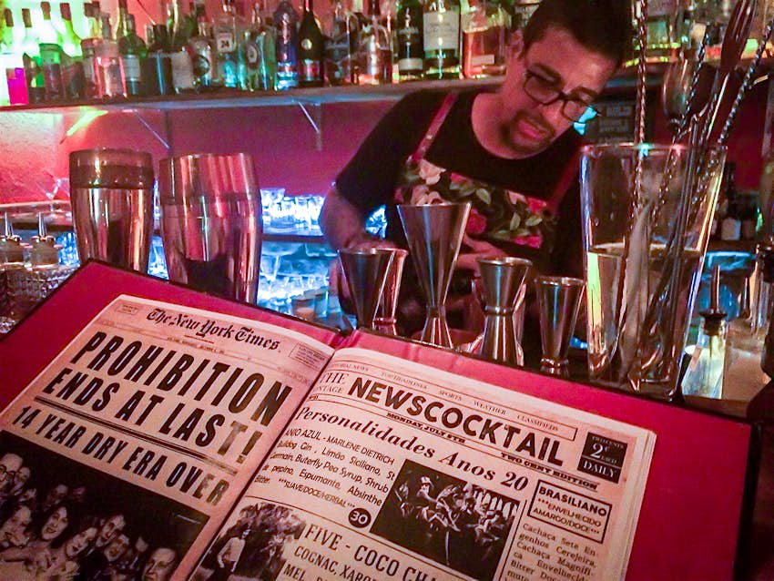 A book with images of Prohibition-era newspapers sit in the foreground as a man with glasses mixes a cocktail in the background