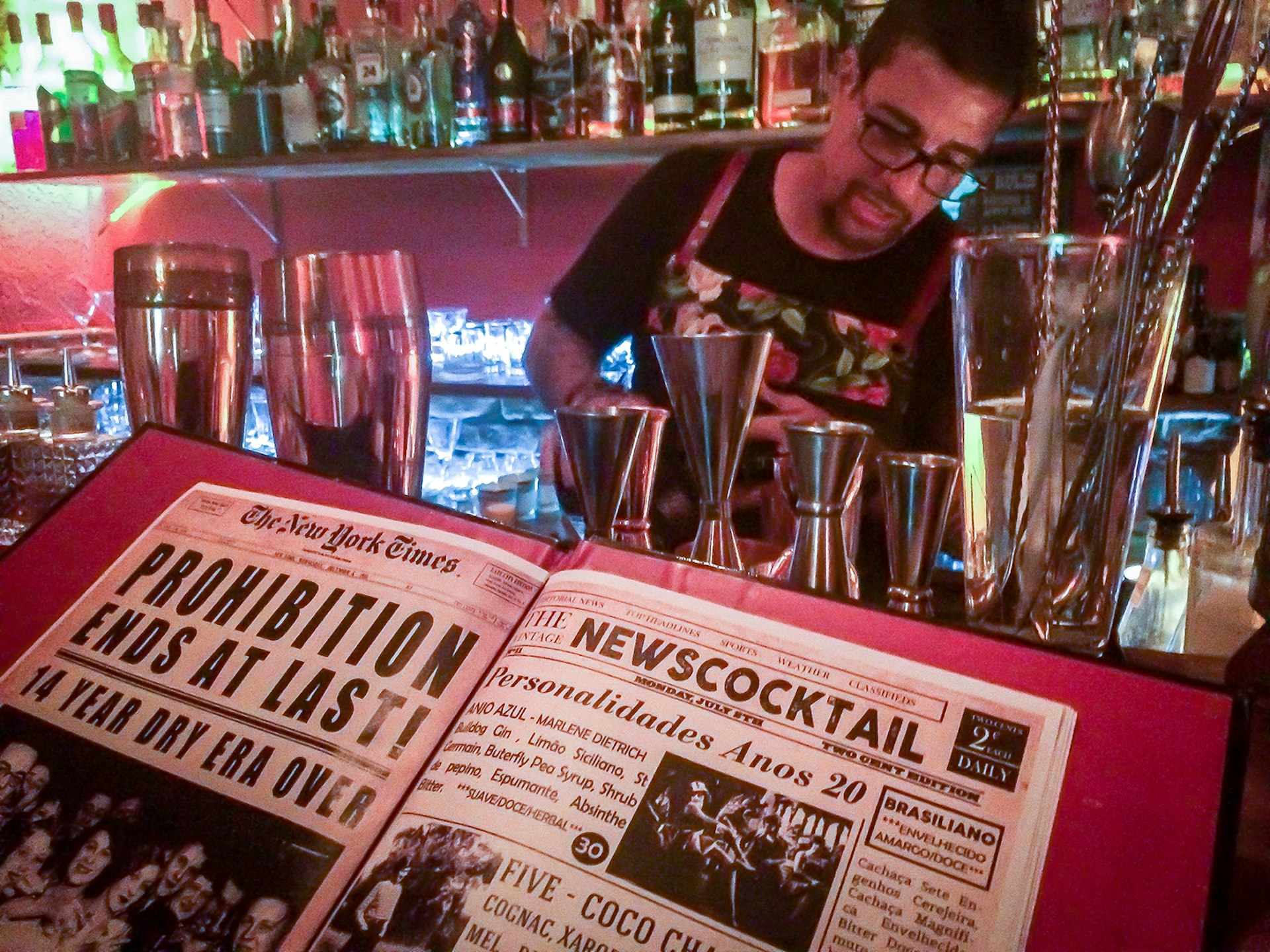 A book with images of Prohibition-era newspapers sit in the foreground as a man with glasses mixes a cocktail in the background