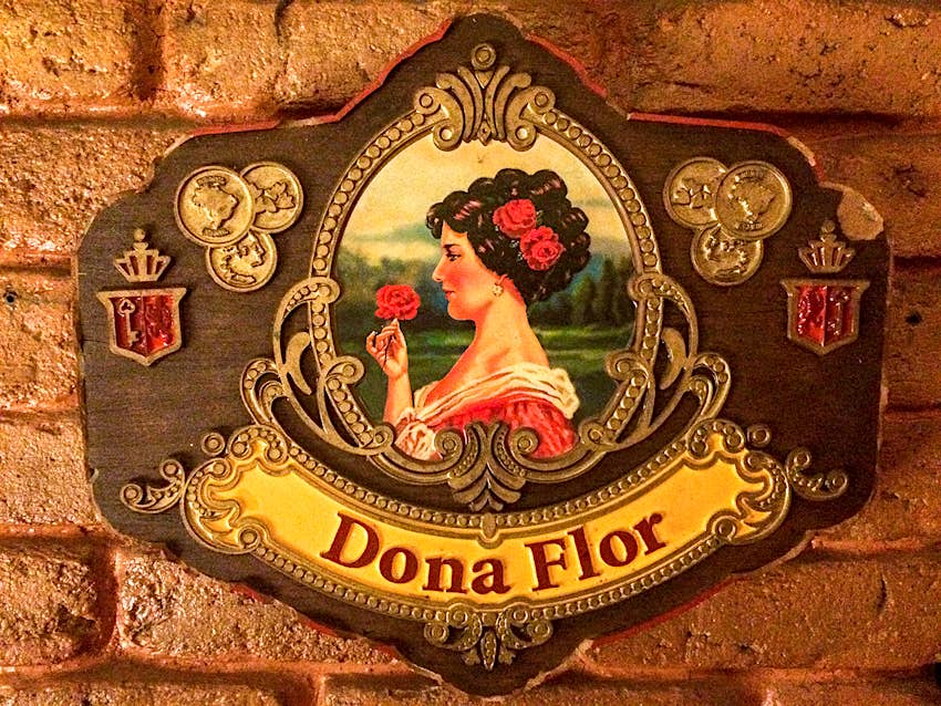 A close-up of a plaque with a painting of a woman with dark hair smelling a pink rose and wearing a pink dress, with "Dona Flor" written below it