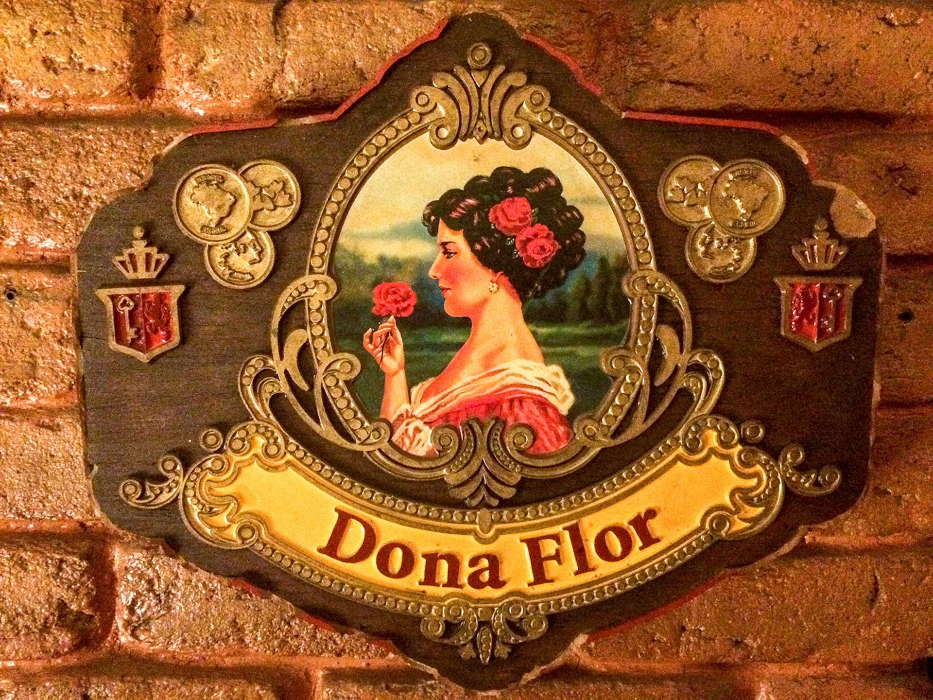 A close-up of a plaque with a painting of a woman with dark hair smelling a pink rose and wearing a pink dress, with "Dona Flor" written below it