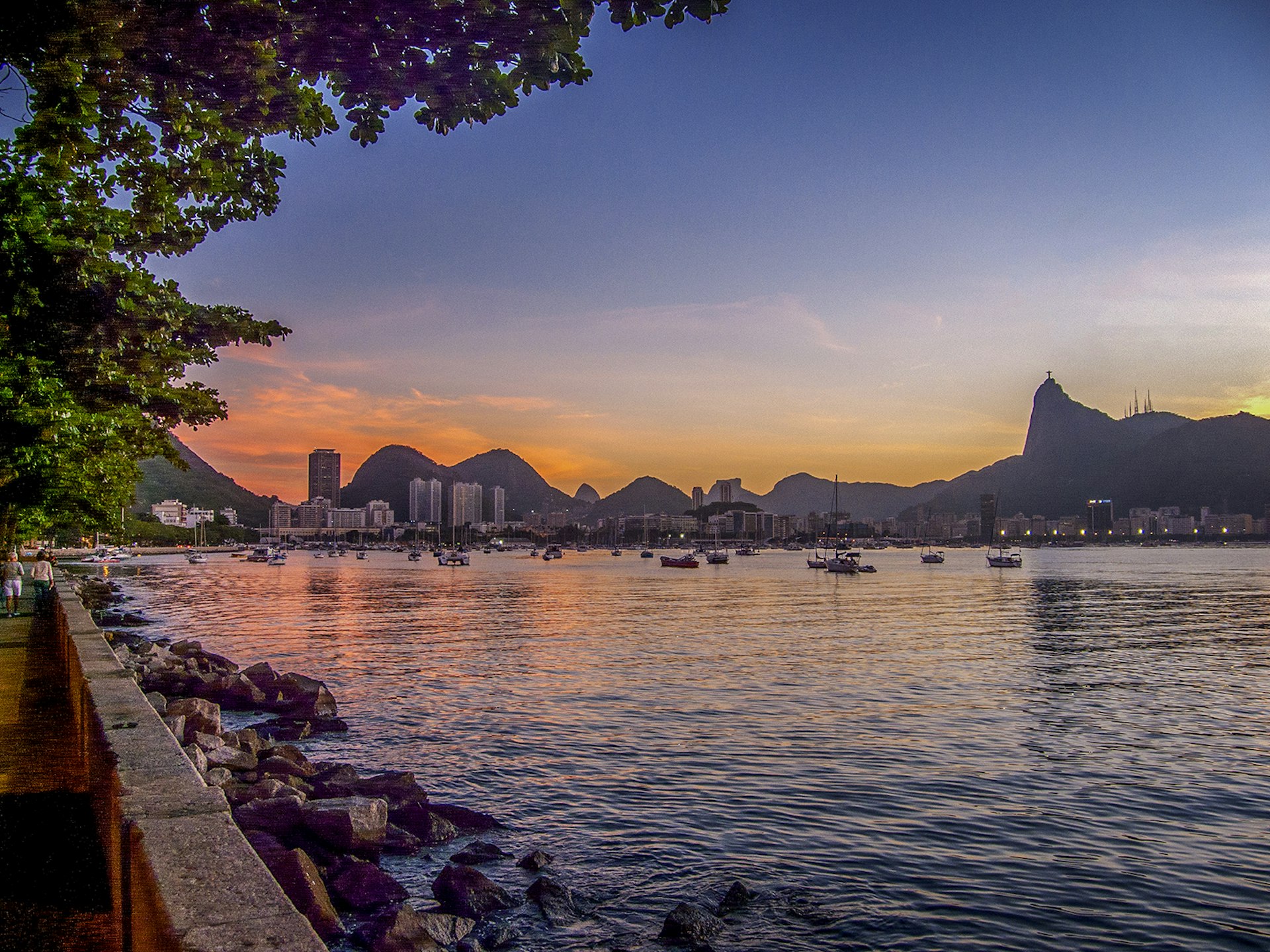 A view of Rio at sunset from a sea wall lining a walking path to the left side of the image, with boats in Guanabara Bay