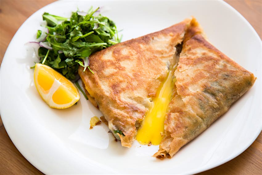Features - brik, egg and tuna turnover, tunisian food Brik, a classic Tunisian dish made with fried pastry and egg. is on a white plate and garnished with a lemon wedge and a pile of green parsley