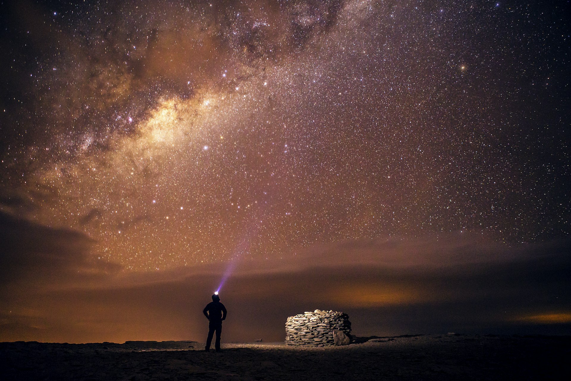 A person stands in the dark next to a stone structure wearing a headlamp; the skies above reveal the Milky Way. Atacama Desert, Chile, South America. 