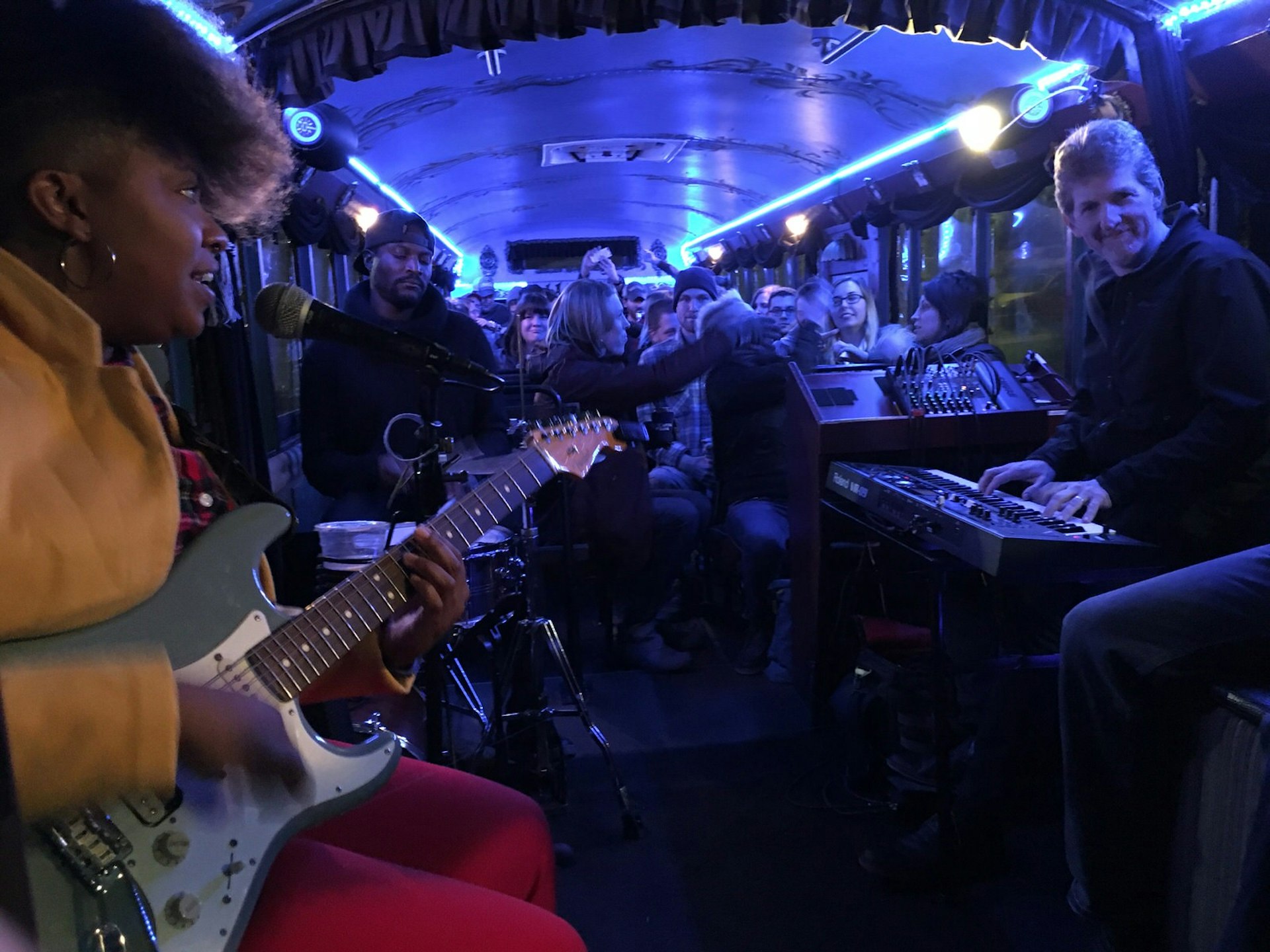 A band, including a woman with electric guitar and microphone, a smiling male keyboardist and a drummer with his eyes closed in concentration, are playing their instruments inside a down-lit bus. Behind them is the audience, crammed into the bus passenger seats. It's LaZoom Band & Beer Bus Tour, a scene in musical city Asheville, North Carolina.