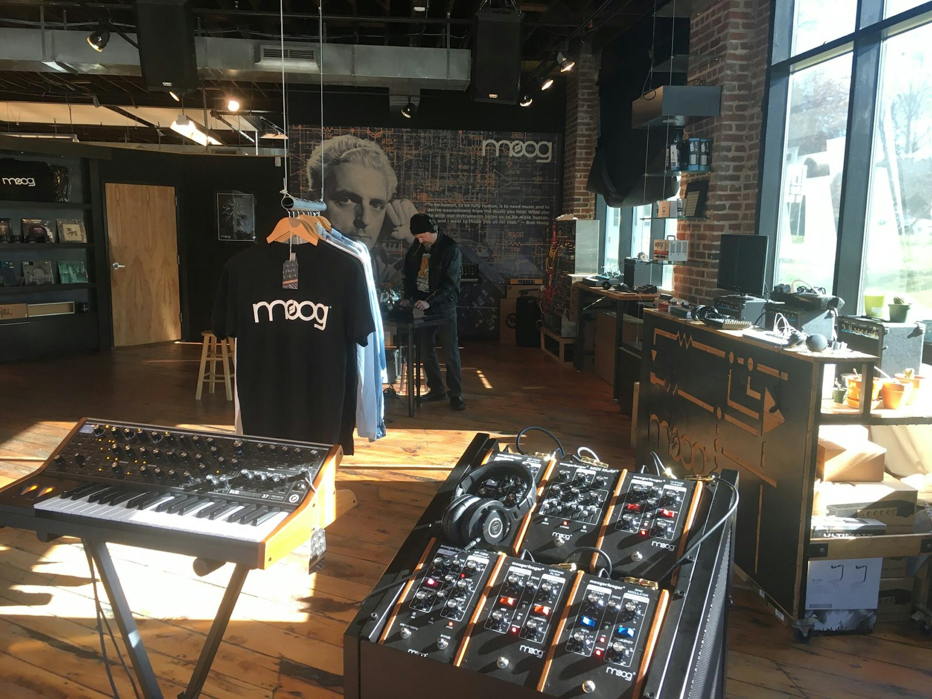 Behind a synthesizer and electronic keyboard are racks of merchandise marked with the logo for 'Moog', an Asheville-born factory that makes electronic synthesizers.