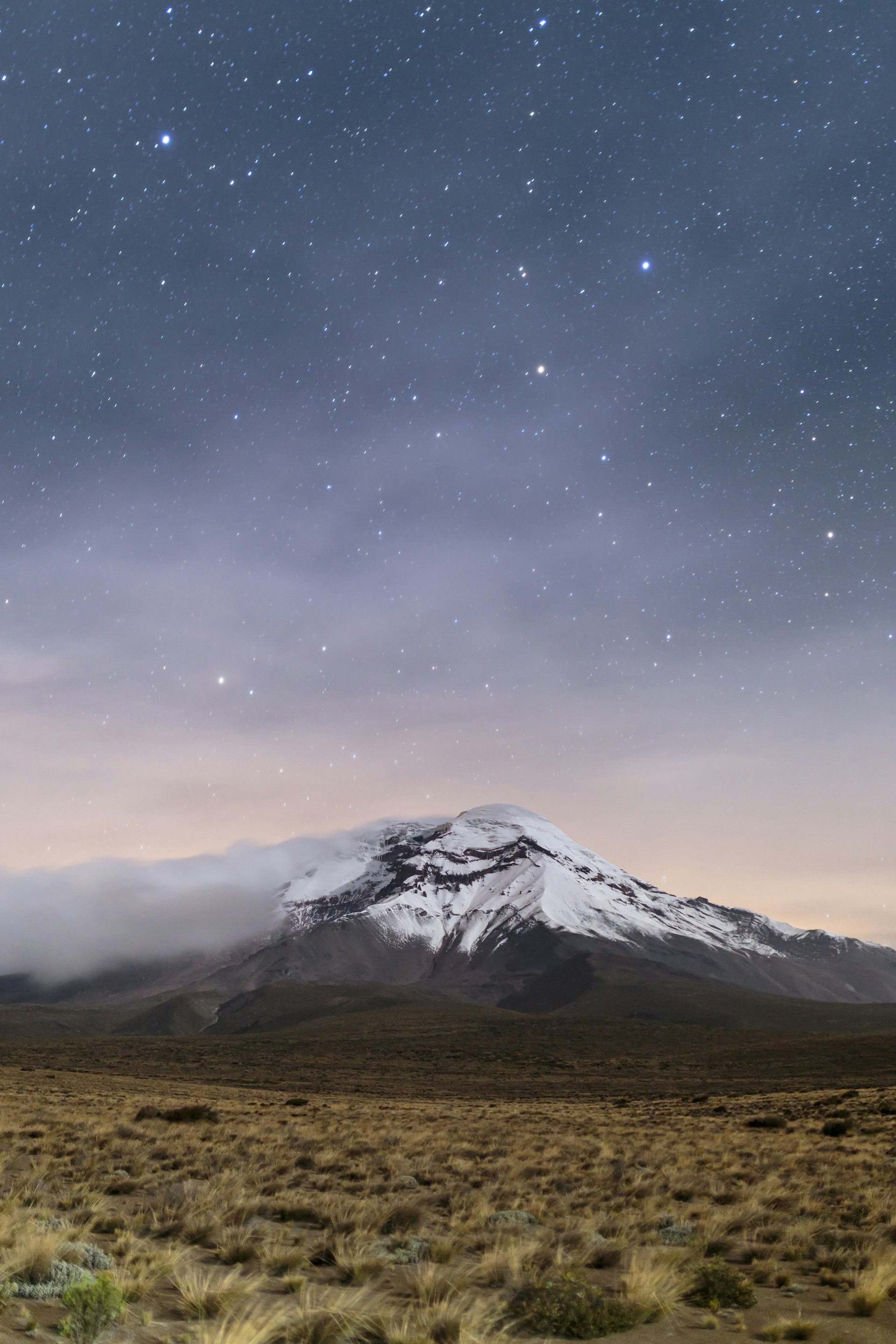 A snowcapped mountain sits at the edge of a grassland in Ecuador; starts permeate the clouds above. 