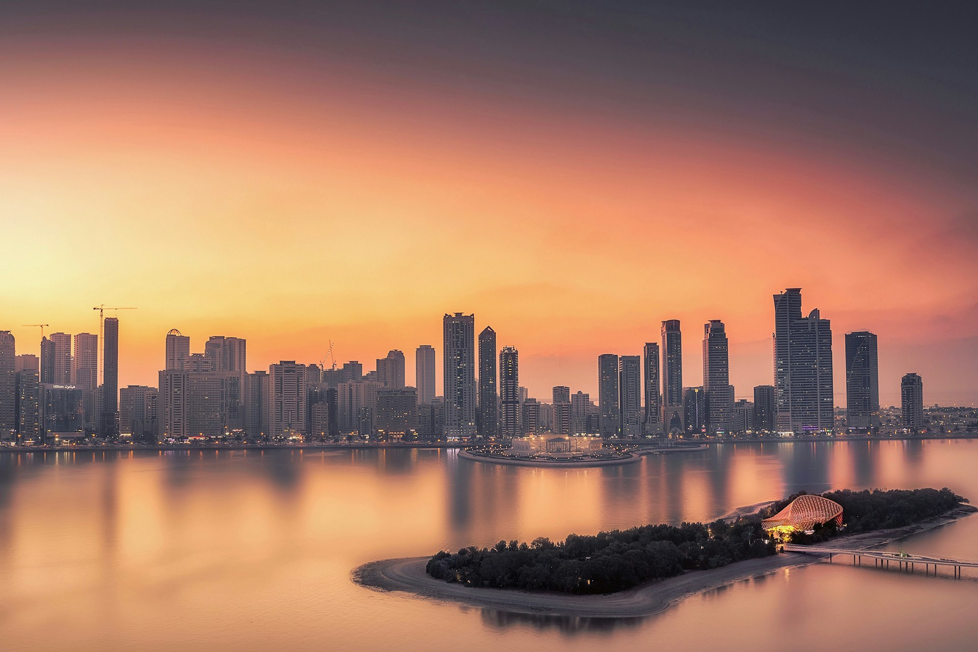 The skyline of Sharjah, comprising numerous skyscrapers, juts up into a bright orange sky at sunset; in the foreground is the forest-covered island of Al Noor, which has some dazzling architecture at its centre.