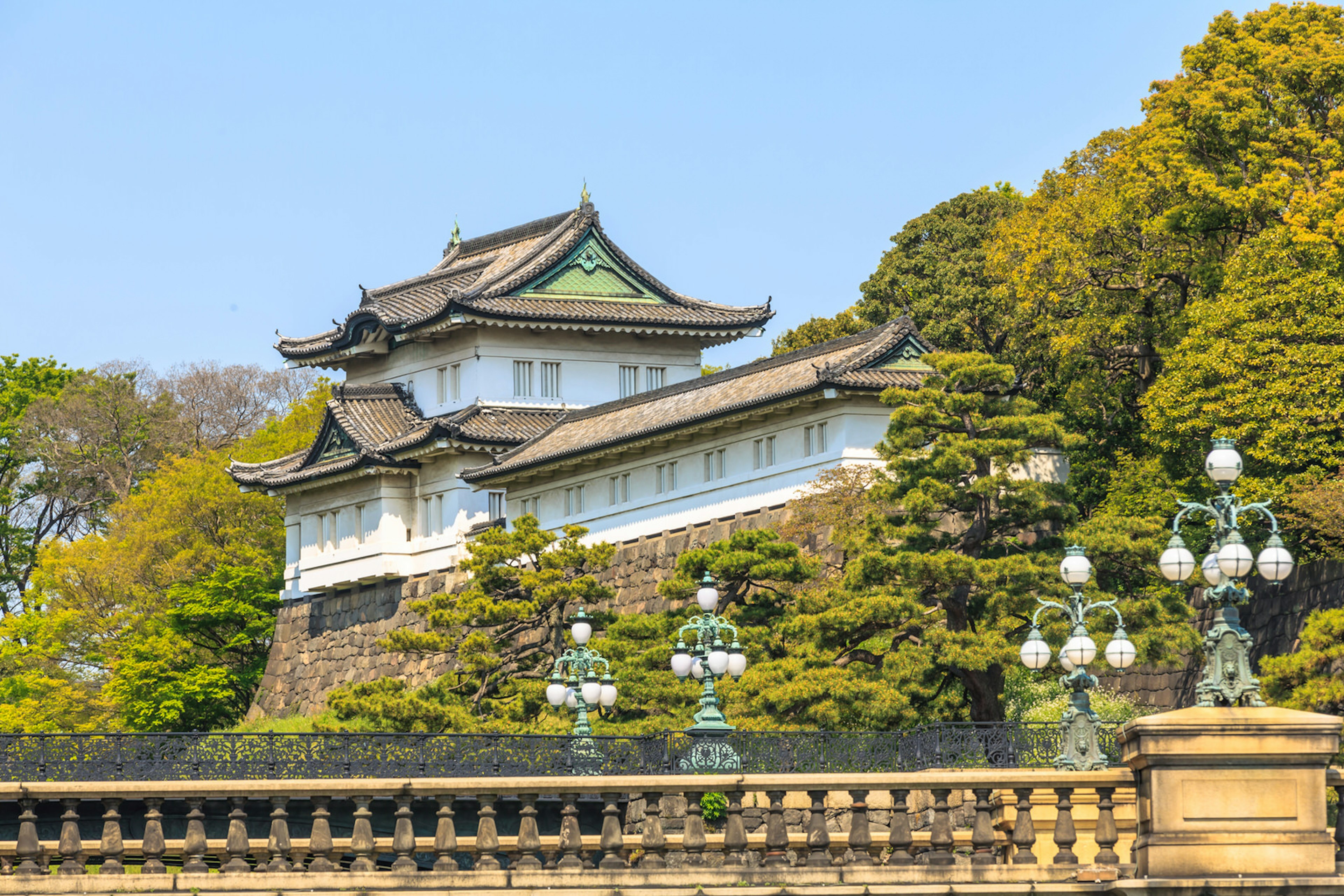 With trees on either side and a balustrade in front, the Imperial Palace in Tokyo, Japan, stands beneath a blue sky, painted white with ornate eaves