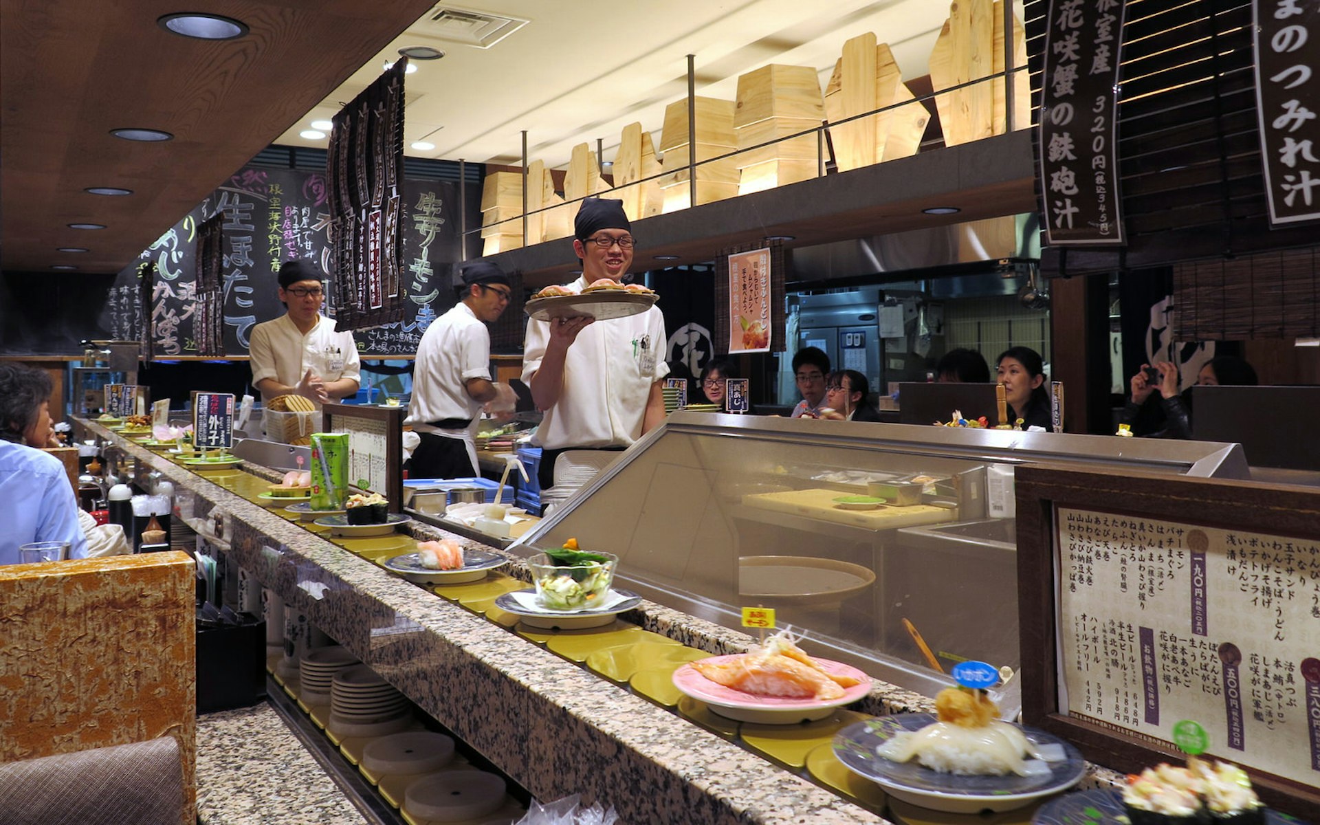A Tokyo restaurant interior with a conveyor belt, upon which are platefuls of sushi and assorted Japanese fare. Behind the belt in the kitchen, chefs clad in white, smiling, prepare and hold aloft dishes.