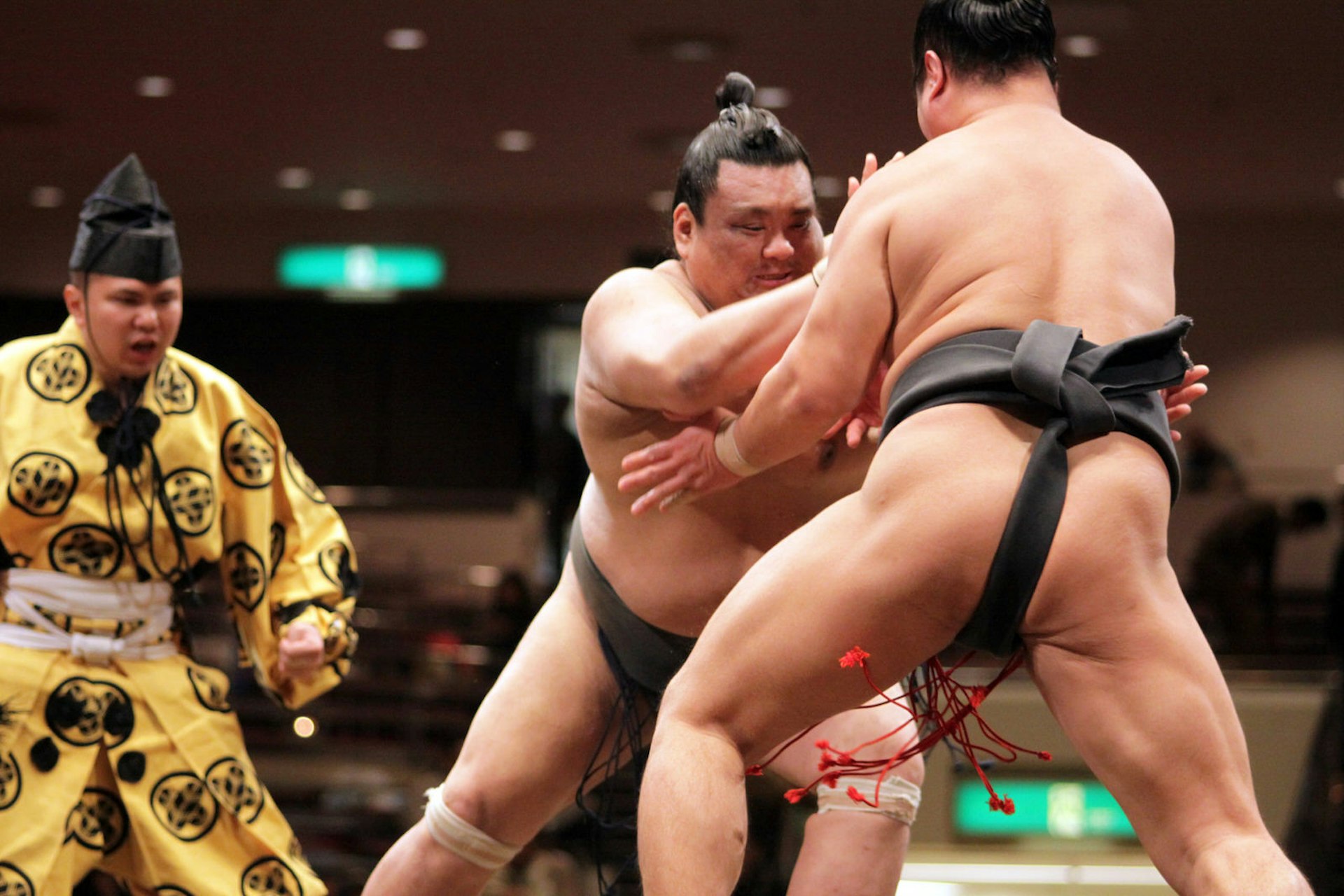 Two sumo wrestlers, their hair tied in top knots, grip one another in a standoff as the referee, clad in a yellow robe, oversees them, at the Tokyo Grand Sumo Tournament, Japan