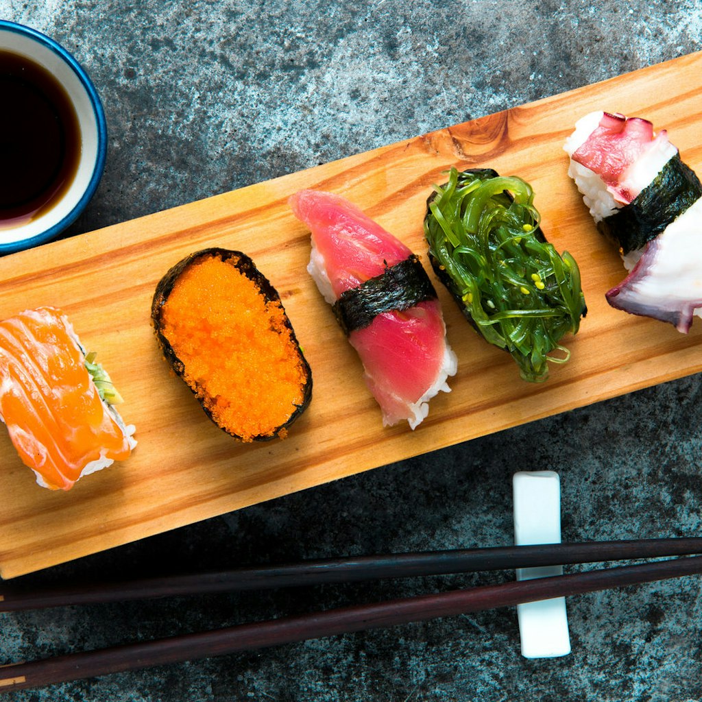 Seen from above, a wooden board with a selection of sushi arranged across it -- topped with fish roe, seaweed and thinly sliced fish. Chopsticks are laid next to the board, as is a small bowl of dark soy sauce, which contrasts with a blue speckled tabletop at a Tokyo restaurant.