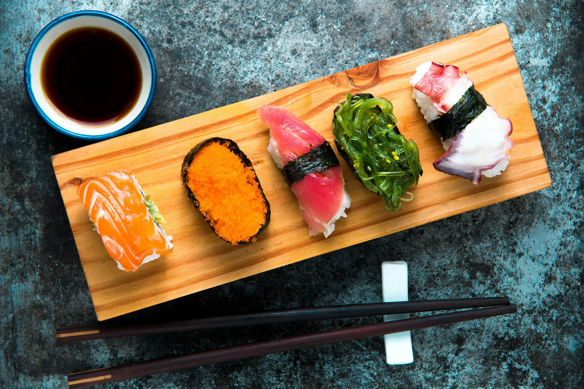 Seen from above, a wooden board with a selection of sushi arranged across it, topped with fish roe, seaweed and thinly sliced fish. Chopsticks are laid next to the board, as is a small bowl of dark soy sauce, which contrasts with a blue speckled tabletop at a Tokyo restaurant.