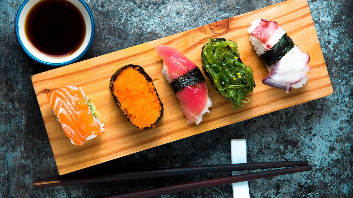 Seen from above, a wooden board with a selection of sushi arranged across it -- topped with fish roe, seaweed and thinly sliced fish. Chopsticks are laid next to the board, as is a small bowl of dark soy sauce, which contrasts with a blue speckled tabletop at a Tokyo restaurant.