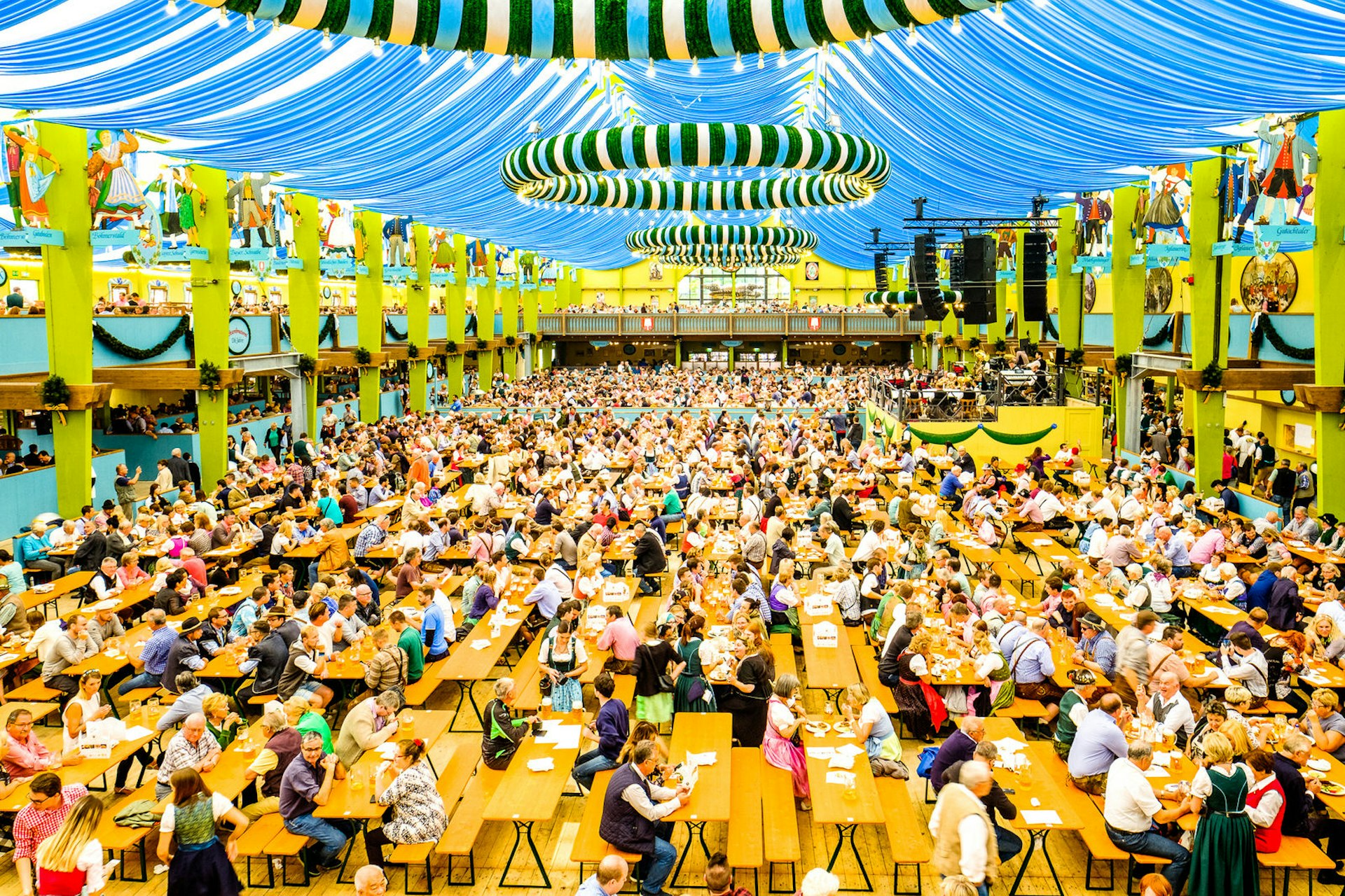 People in the 'Spaten' beer tent at Oktoberfest in Munich. The huge tent, that houses hundreds of tables, has a colourful blue ceiling and striped decorations.