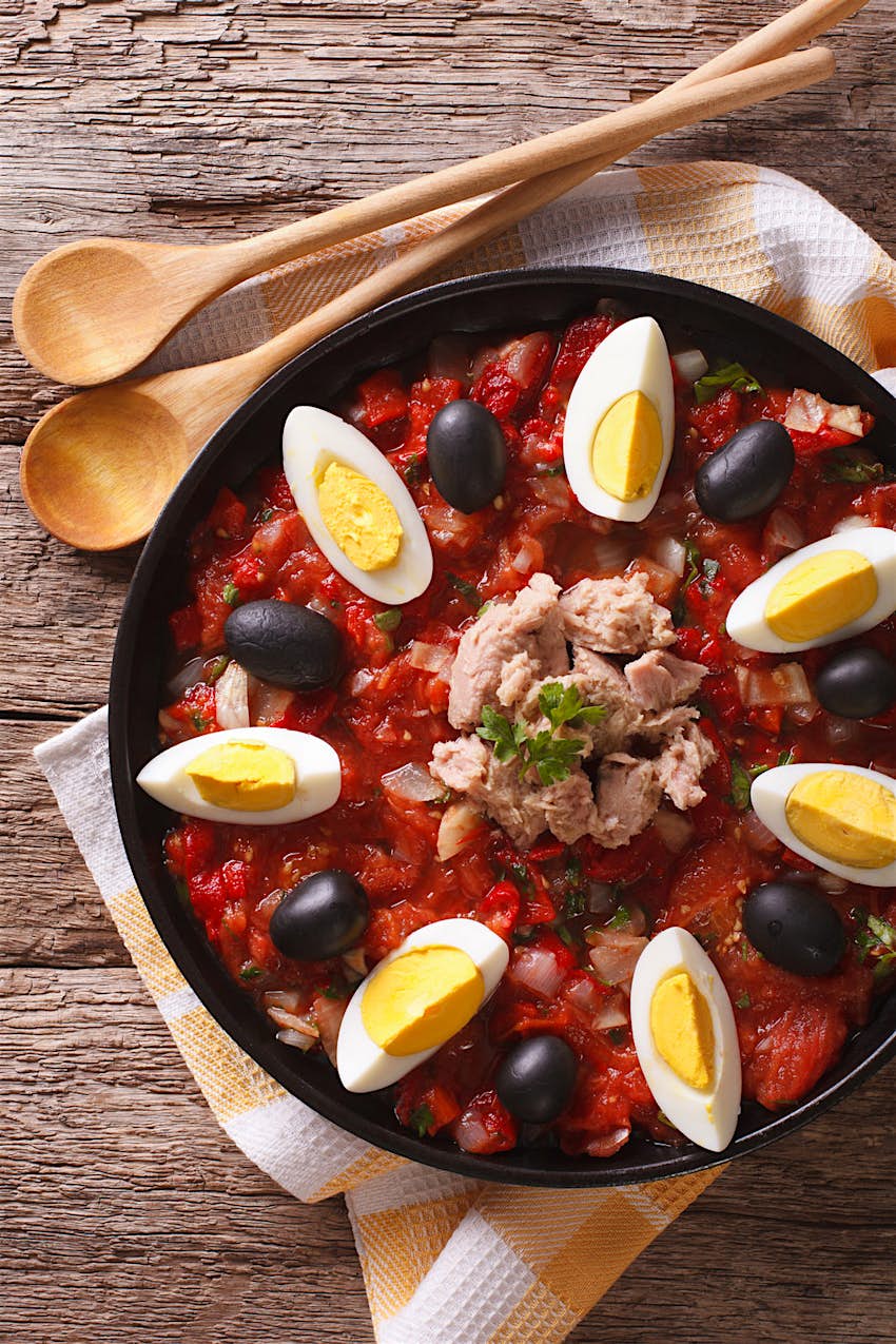 Features - Arabic Mechouia salad with vegetables, tuna and eggs closeup Mechouia salad (slata mechouia) with vegetables, tuna and eggs in a black bowl on a wooden table