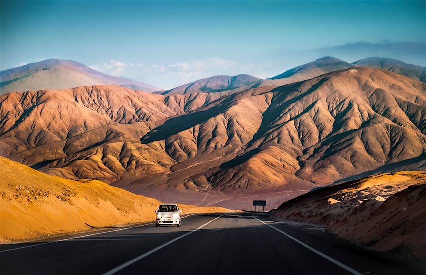A white car drives on the Panamerican Highway through the Atacama desert away from a brown, sandy-looking mountain range