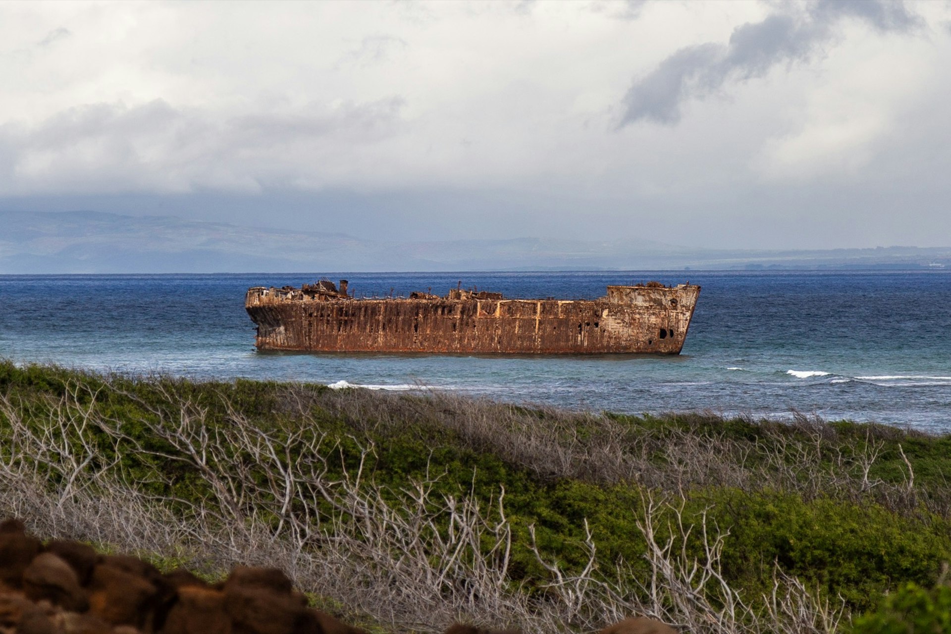 A rusted hull of a huge ship is seen just off the coast of an island, as waves crash between it and the shore and scrub brush fills the foreground