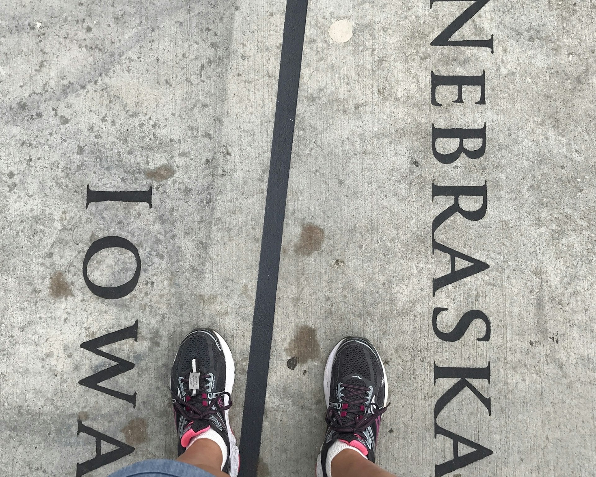 A top-down look at two feet standing on concrete, straddling a line labeled Iowa on the left and Nebraska on the right