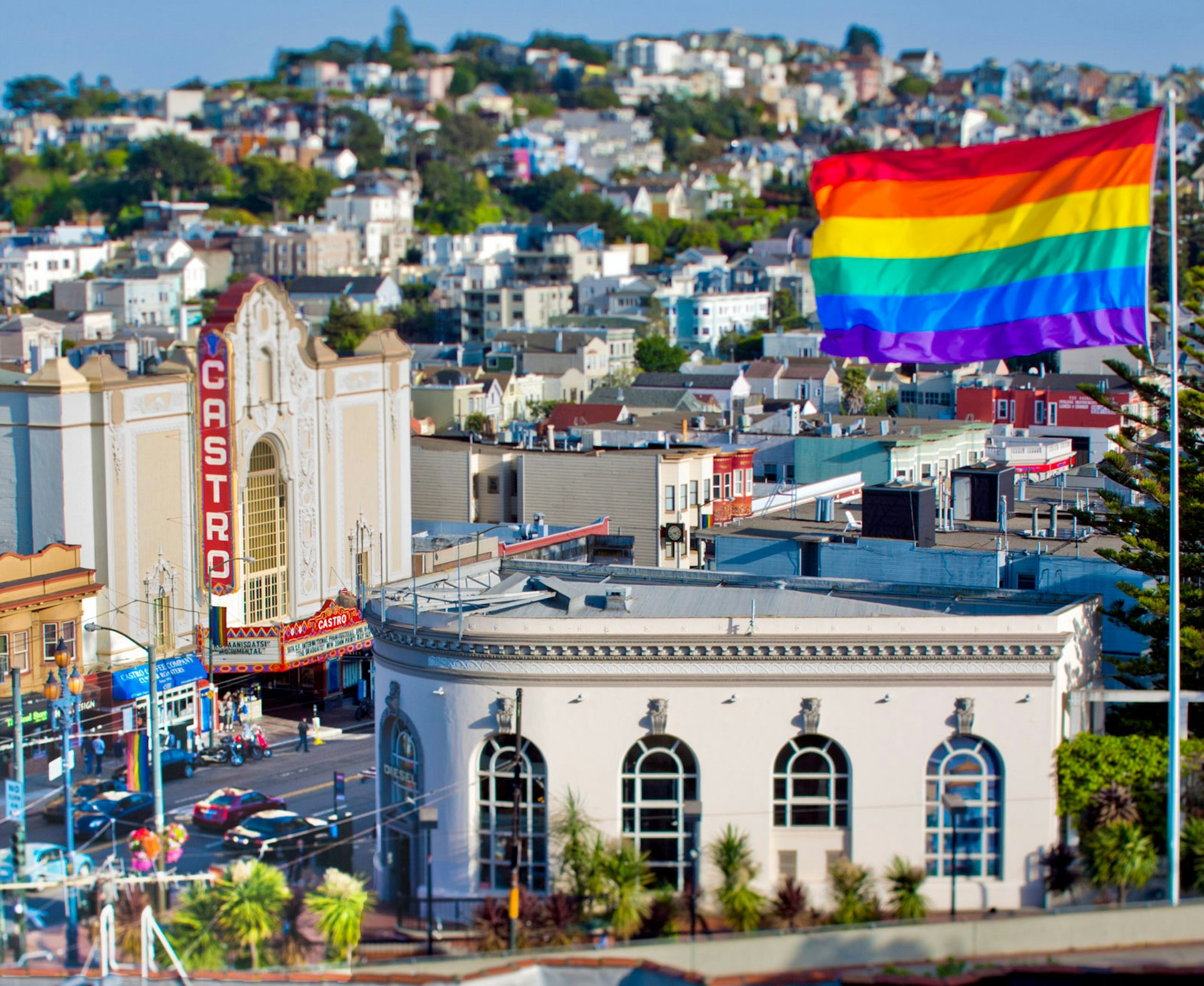 A rainbow flag flies over a white building with the Castro theater in the background and houses creeping up the hill