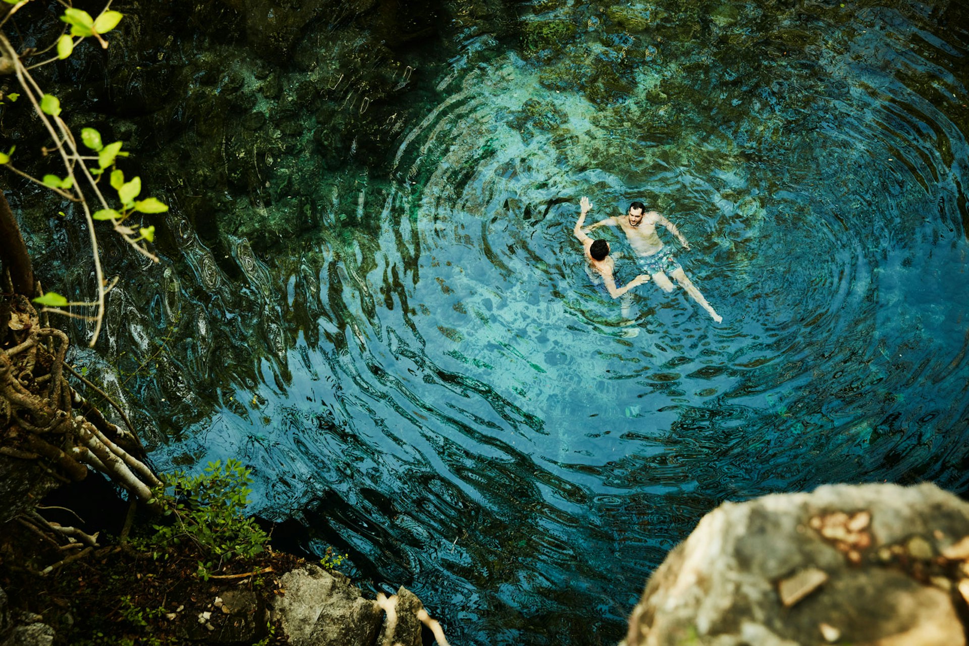 Two men talk and swim in a clear lake with jungle plants and rocks surrounding them