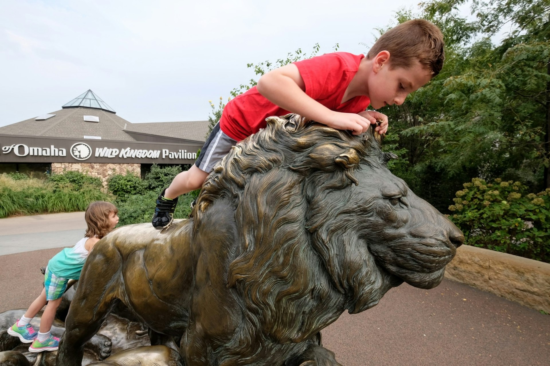 A young boy and a young girl climb on a bronze statue of a lion outside the Omaha Zoo